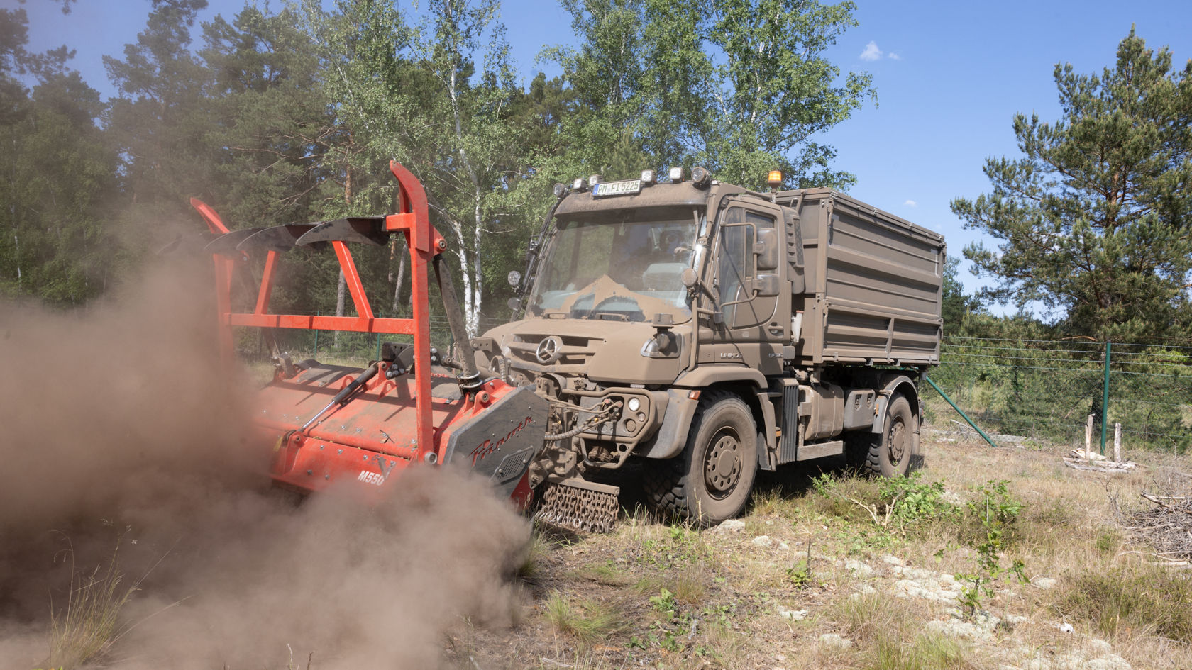 A grey Unimog with a crane body parked in a square. The crane picks up branches and twigs and feeds them into a shredder, which blows the shredded material into a large blue container. There are more branches and twigs lying around the truck.