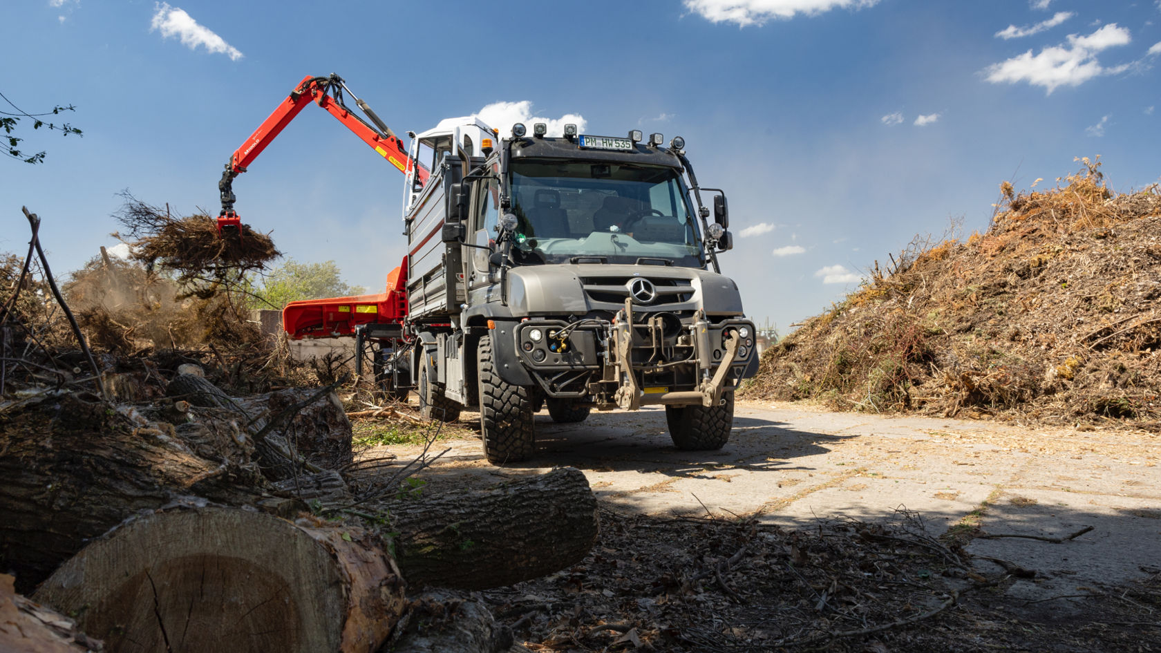 A grey Unimog with a red crane and tip box collecting branches and tree cuttings in a yard with tree trunks and wood waste. The crane is lifting the branches and loading them onto the loading area. Branches and tree parts are piled up around the vehicle. 