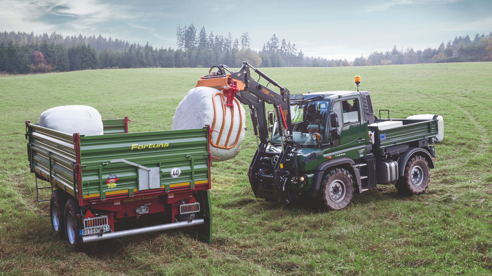 A green Unimog standing in a meadow. Using a crane arm, it is lifting a large white hay bale onto a trailer in front of it. A forest can be seen in the background.