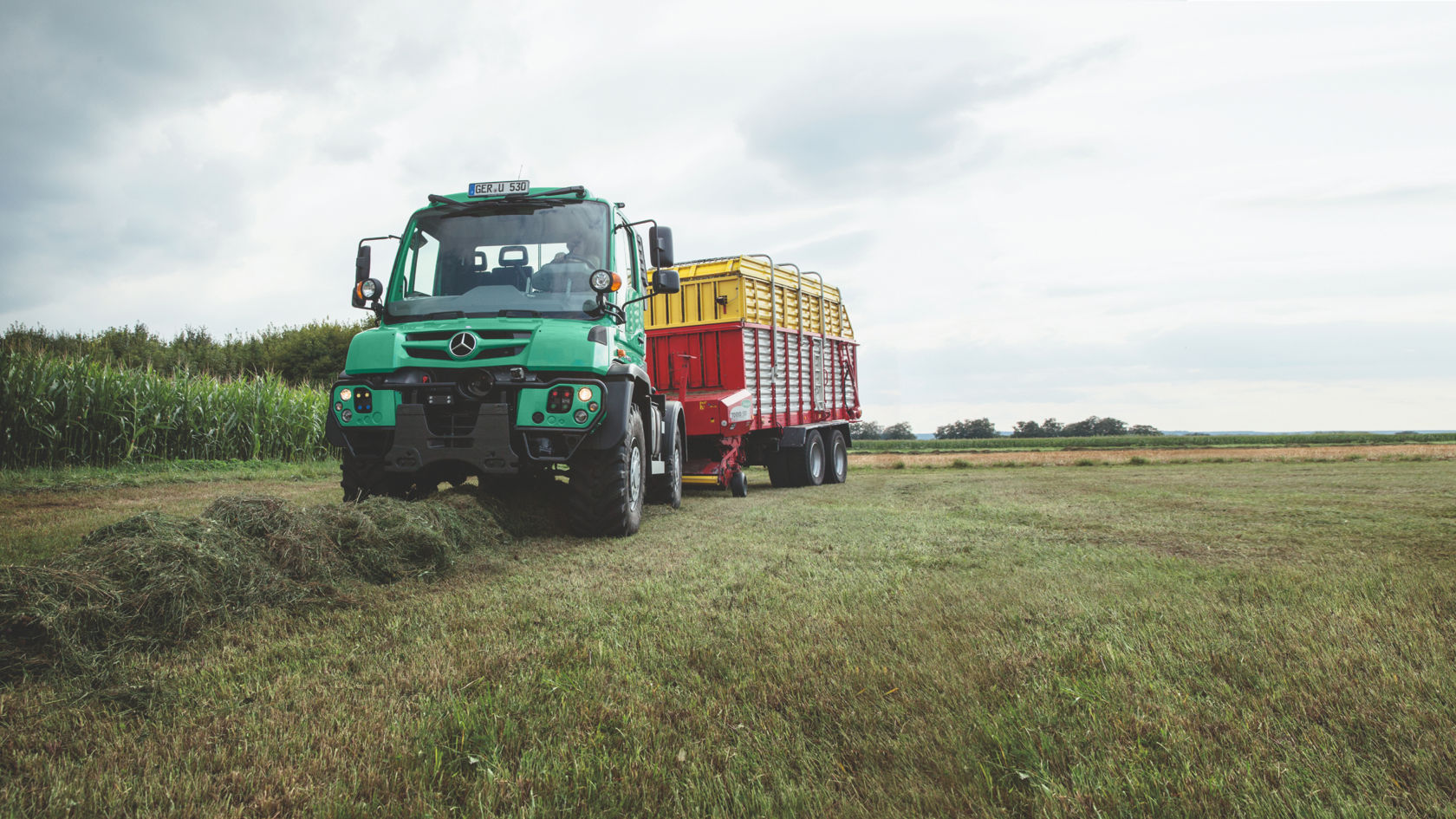 A green Unimog standing in a freshly mowed meadow. The cuttings are in front of it. It is pulling a red and yellow trailer with a latticed body. In the background is a field of corn with trees on the horizon.