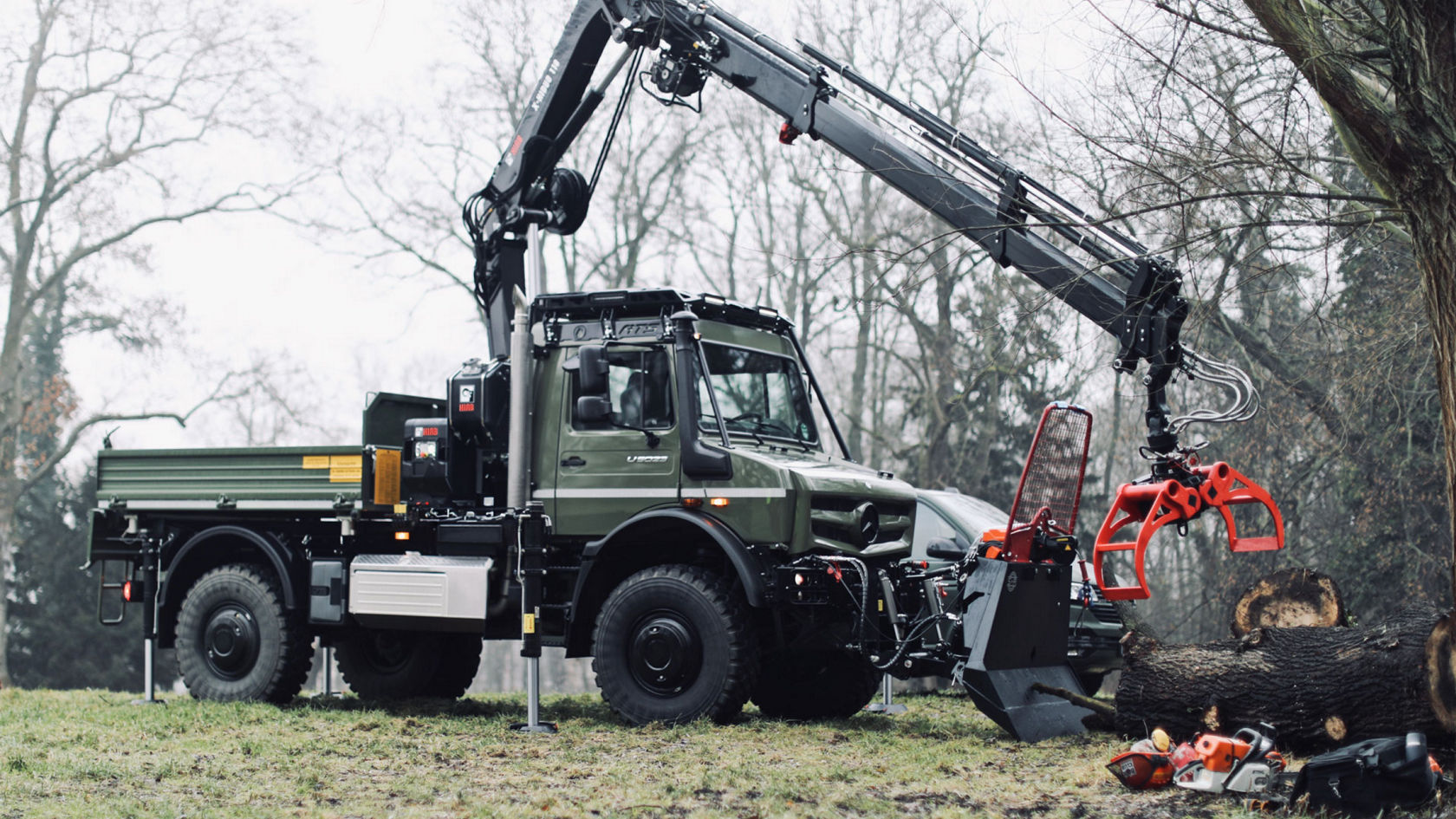 Na obrázku je šedé vozidlo Unimog jedoucí po silnici mezi budovami. Na ložné ploše nákladního vozidla se nachází žluté rýpadlo. Nákladní vozidlo je opatřeno nápisem „Podpora při katastrofách“. Budovy v pozadí jsou tradiční německé hrázděné domy s barevnými okenicemi.