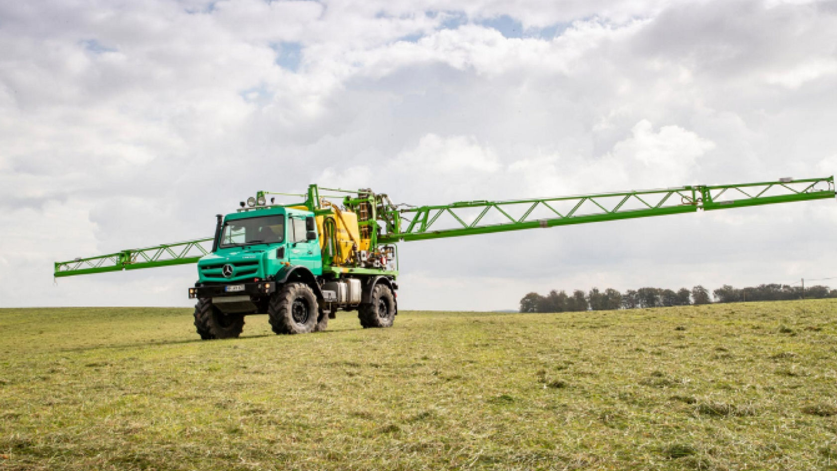 A turquoise Unimog with a long, extended spraying rod standing on a field. The green rod is leaning out far beyond the driver’s cab. A row of trees can be seen in the background.