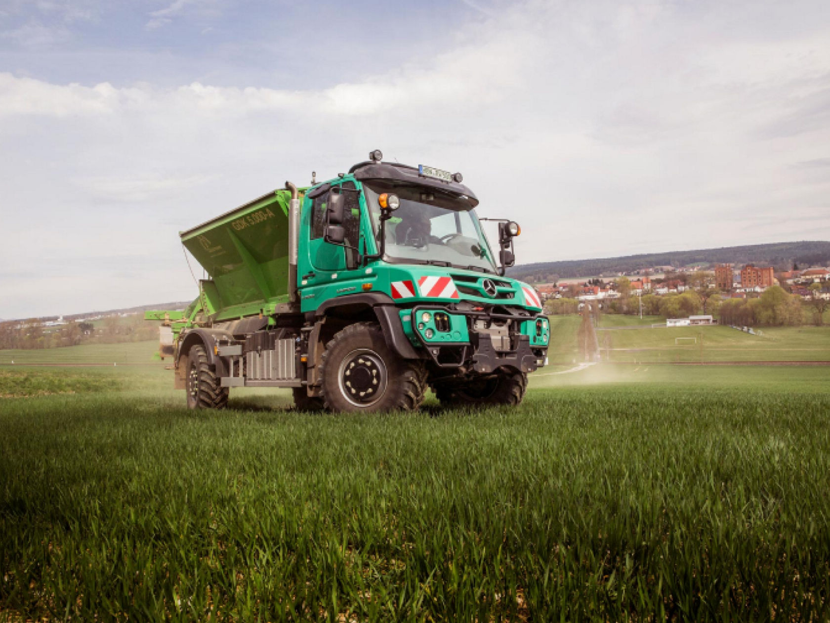 A turquoise Unimog with a long, extended spraying rod standing on a field. The green rod is leaning out far beyond the driver’s cab. A row of trees can be seen in the background.