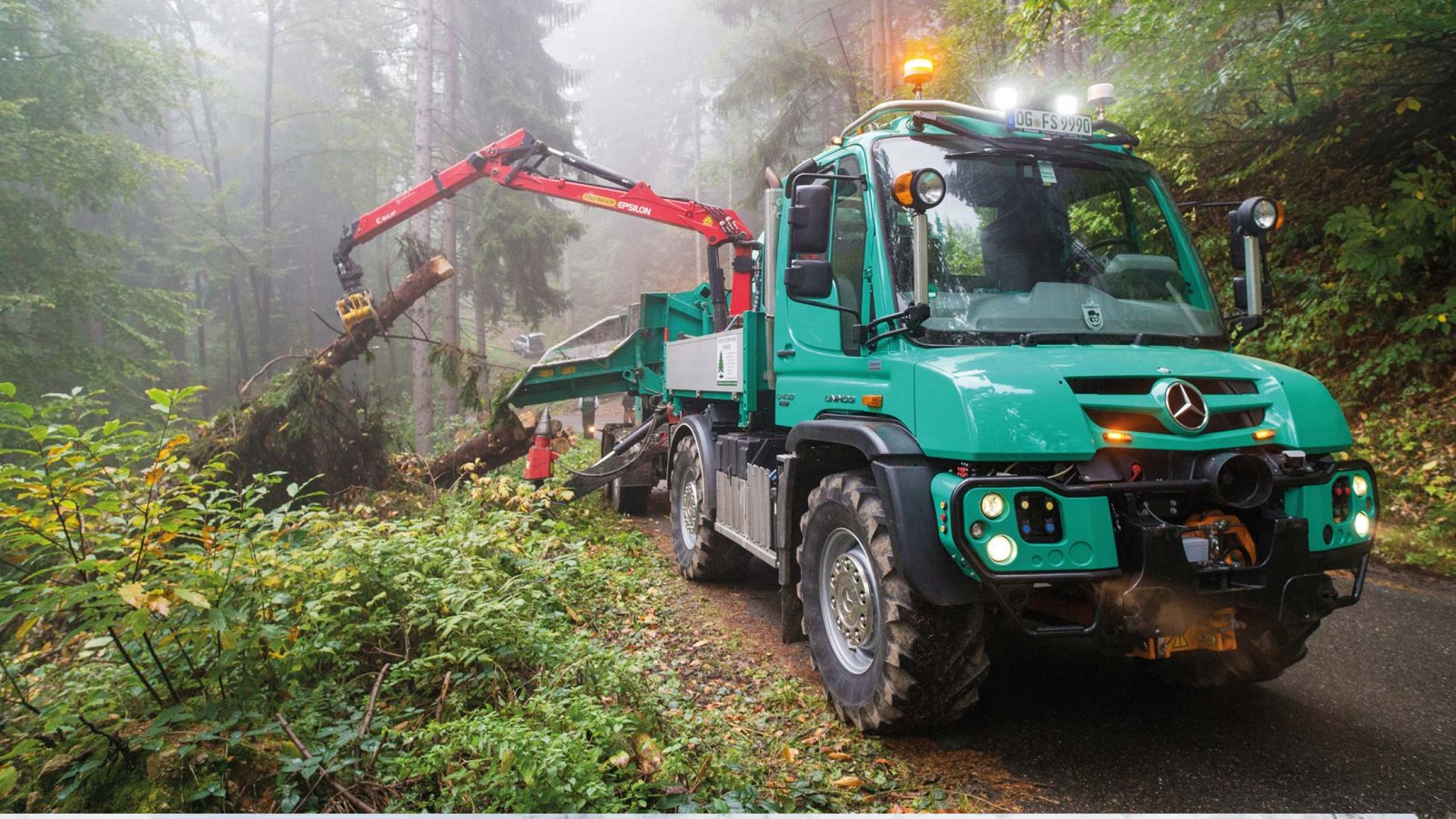 A turquoise Unimog standing on a forest path. Using a red crane arm, it is lifting a tree trunk onto the loading area. The headlights are on because it is foggy in the forest and visibility is poor.