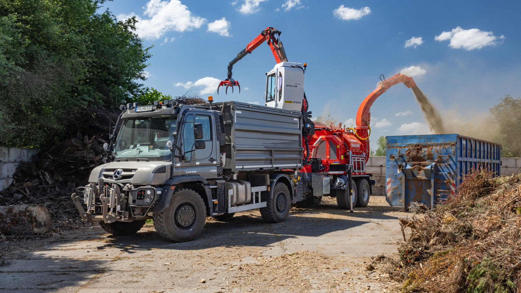 A grey Unimog with a crane body parked in a square. The crane picks up branches and twigs and feeds them into a shredder, which blows the shredded material into a large blue container. There are more branches and twigs lying around the truck.