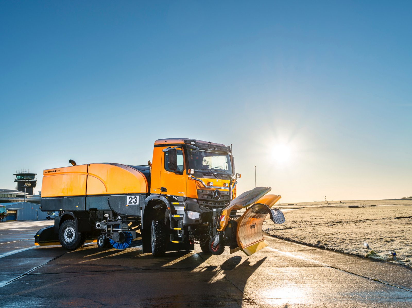 La imagen muestra un Mercedes-Benz Trucks Arocs naranja con pala quitanieves y barredera en un aeropuerto. En segundo plano se ven la torre de control y la pista de despegue y aterrizaje. El cielo es azul y el sol brilla.