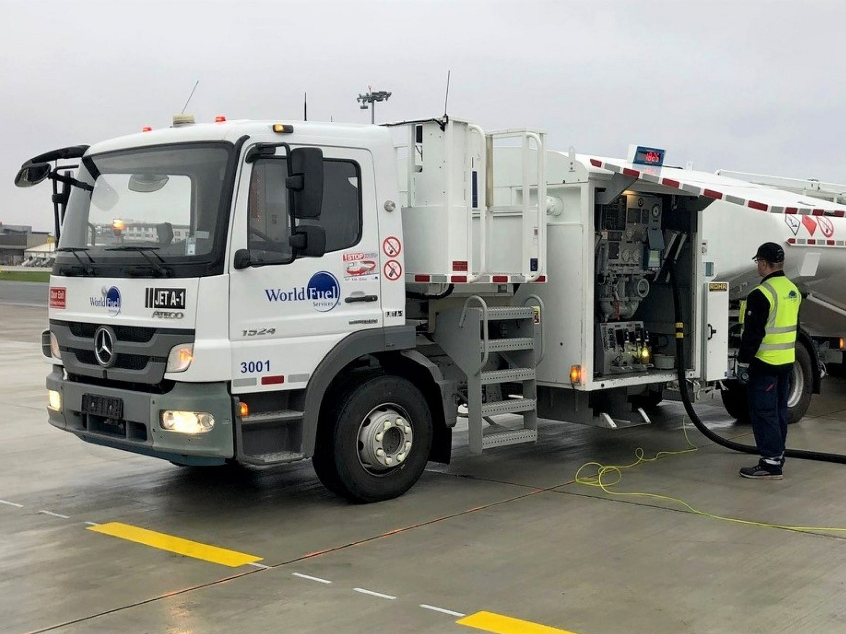 Kuvassa on World Fuel Servicesin valkoinen Mercedes-Benz Trucks Atego -säiliöauto lentokentällä. Työntekijä keltaisessa turvaliivissä seisoo säiliöauton vieressä ja käyttää tankkauspistettä. Taivas on pilvinen.