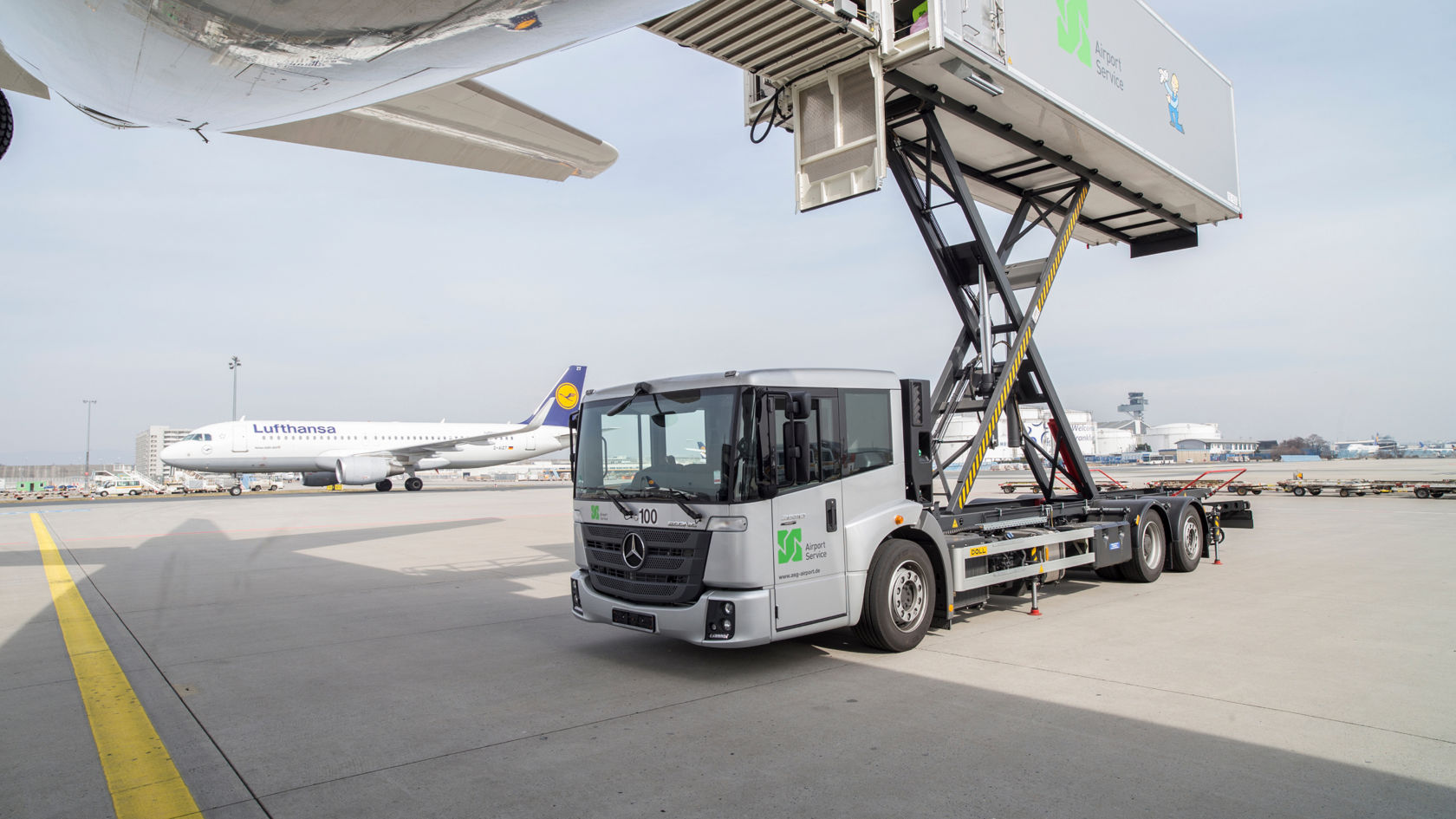 Un Econic plateado con trampilla de carga plegada se encuentra en la plataforma del aeropuerto. La trampilla de carga se encuentra debajo del ala de un avión que no se puede ver. En segundo plano se ve un avión de Lufthansa.