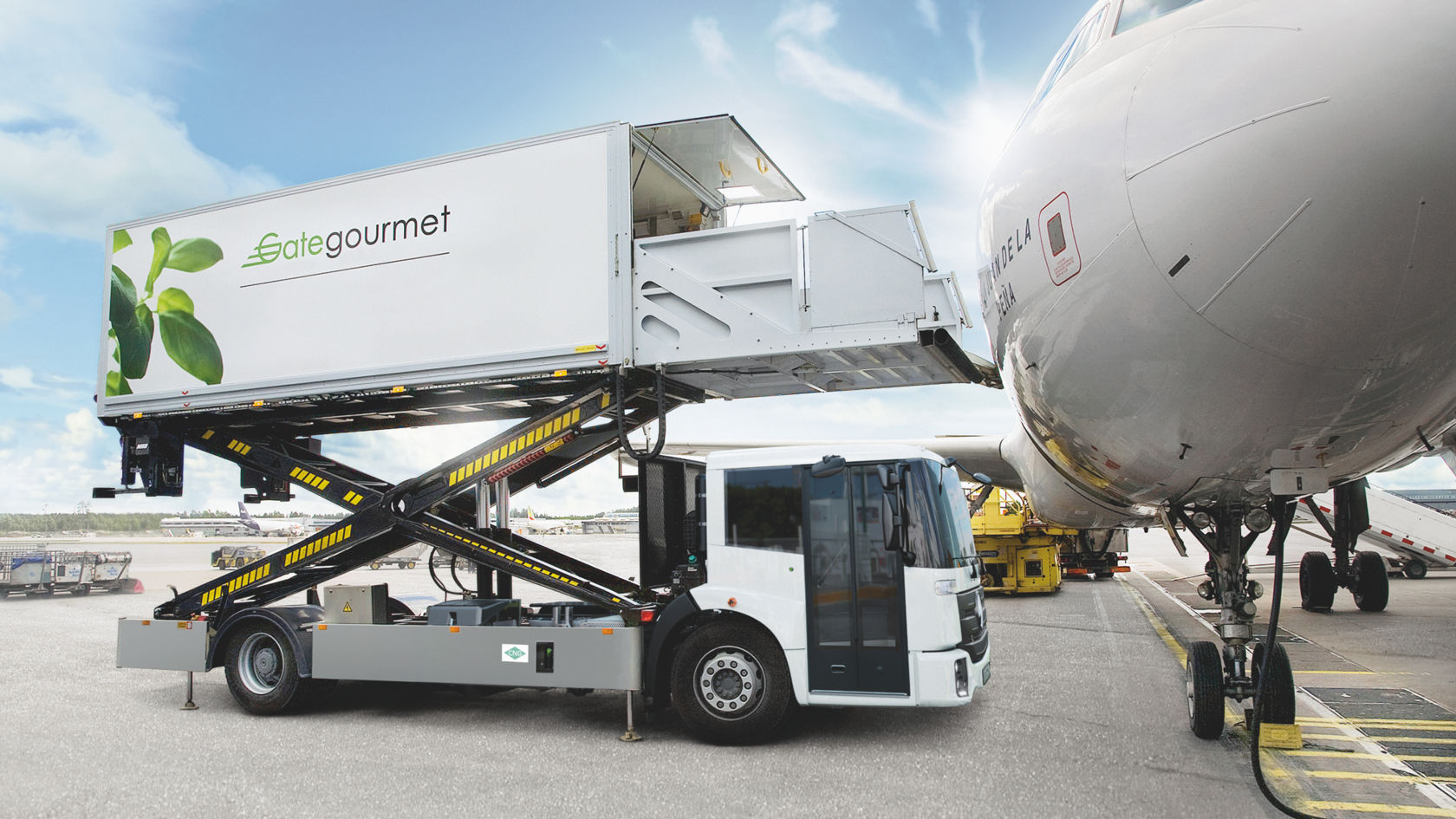 Un Econic blanco y gris con trampilla de carga abatida está aparcado delante de un avión. En la superestructura se lee «Gate Gourmet». La trampilla de carga llega hasta la puerta del avión. En segundo plano se ven otros vehículos y aviones. 
