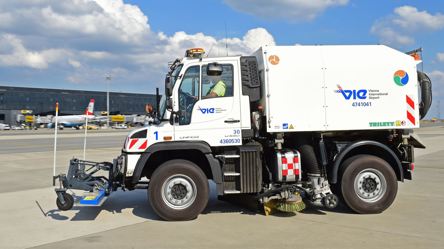 Valkoinen Unimog ajaa lentokentän asematasolla. Lakaisuauton sivulla on suuri päällirakenne, jossa on harjoja ja edessä pienempi luuta. Taustalla näkyy lentokoneita ja lentokenttärakennus. Päällirakenteessa lukee "VIE Vienna International Airport".