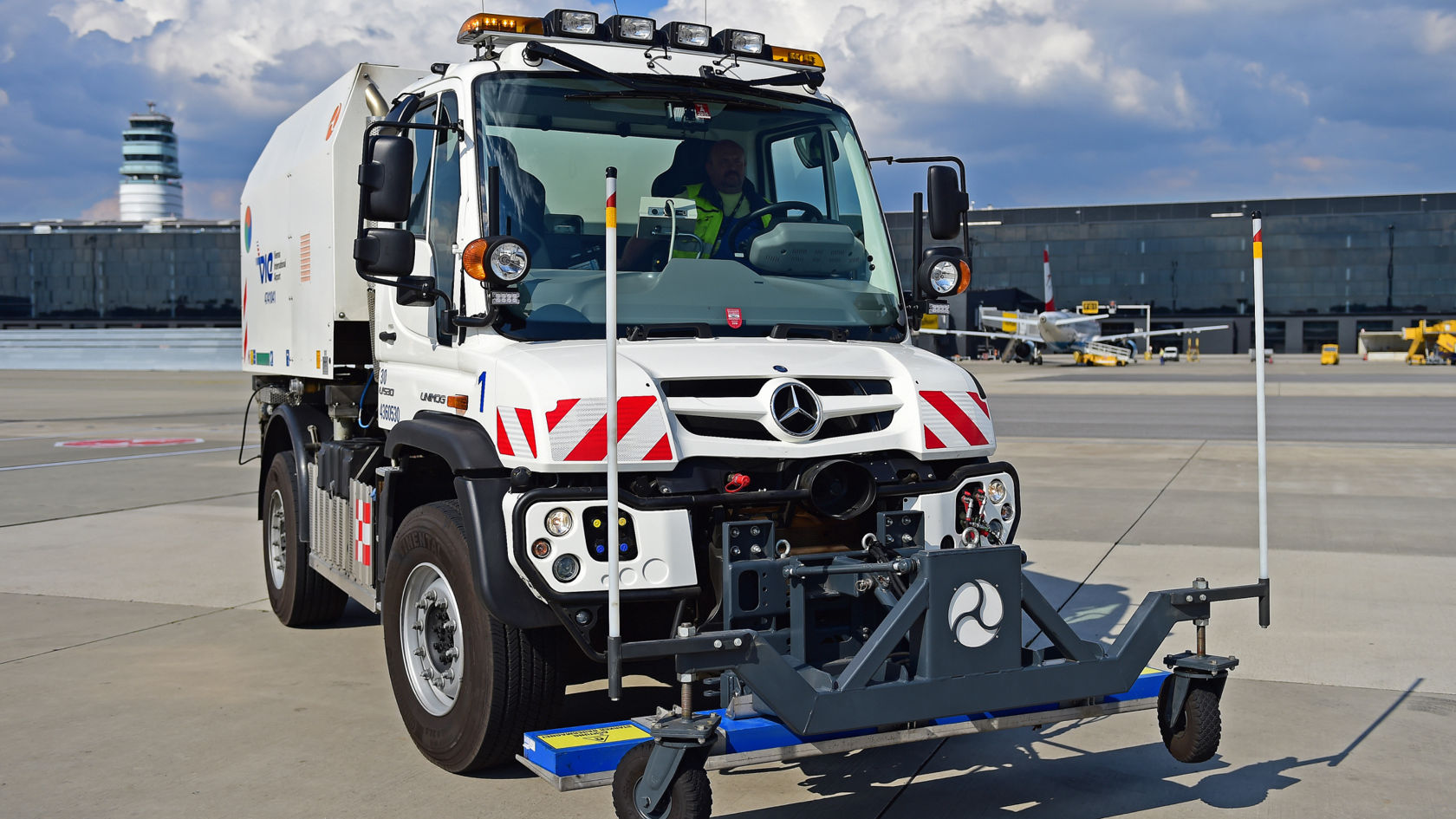 Valkoinen Unimog, jonka etuosassa on puhdistuslaitteisto, seisoo lentokentän asematasolla. Pesulaitteisto koostuu rungosta, jossa on harjoja ja pyöriä. Taustalla näkyy lentokoneita ja torni.