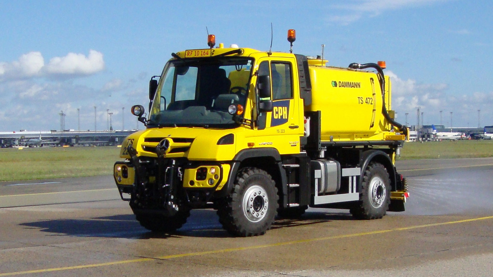 Talvikunnossapitovarustuksella varustettu oranssi Unimog ajaa tiellä. Kuorma-autossa on päällirakenne, jossa on suljettu rullaovi ja ruiskutuspalkki takana. Päällirakenteessa lukee "Winterdienst". Taustalla näkyy lumihuippuisia vuoria.