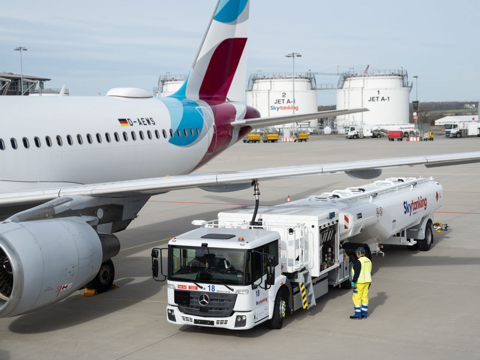 Un Econic blanco con equipamiento para depósitos de combustible está junto a un avión en la plataforma del aeropuerto. En el depósito de combustible se lee «Skytanking». Junto al camión hay un empleado con ropa de alta visibilidad. En segundo plano se ven otros aviones y grandes depósitos de combustible.