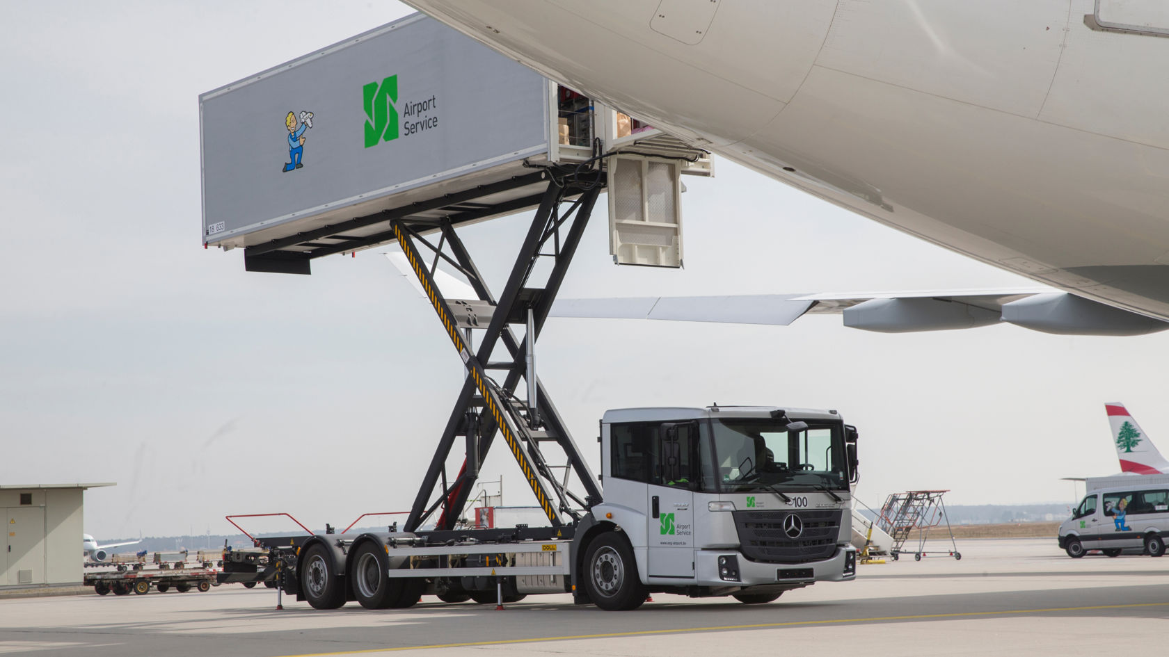 Harmaa Econic, jonka takalaitanostin on käännetty ylös, seisoo lentokentän asematasolla. Takalaitanostin sijaitsee lentokoneen siiven alla. Takalaitanostimessa lukee "Airport Service". Taustalla näkyy muita lentokoneita ja rakennus.