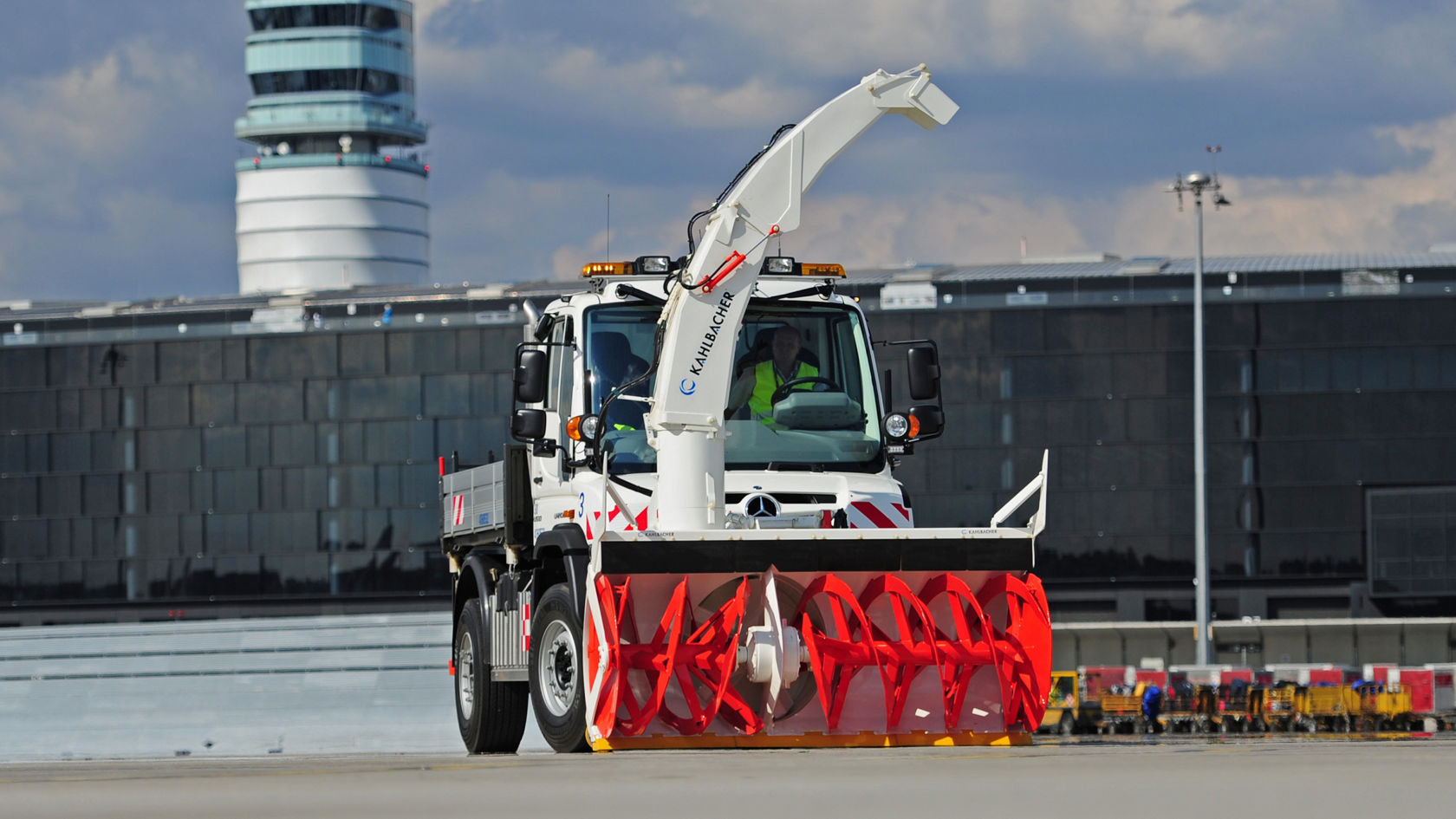 Valkoinen Unimog, jonka edessä on punainen lumilinko, seisoo lentokentän rullaustiellä. Lumilingossa lukee "Aebi Schmidt". Taustalla näkyy torni ja lentokenttärakennus.
