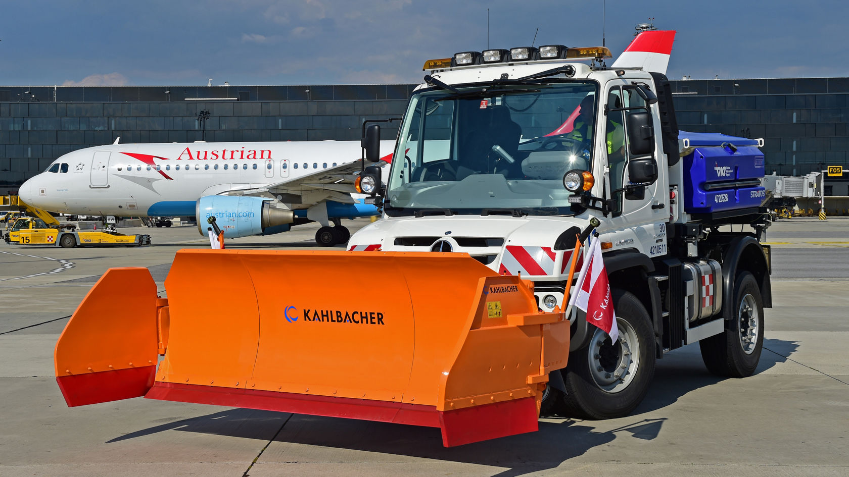 Valkoinen Unimog, jonka edessä on oranssi lumiaura, seisoo lentokentän asematasolla. Lumiaurassa lukee "Kahlbacher". Taustalla näkyy Austrian Airlinesin lentokone. Unimogin sivuille on kiinnitetty punavalkoiset varoitusliput. Kuorma-auton perässä näkyy sininen säiliö.