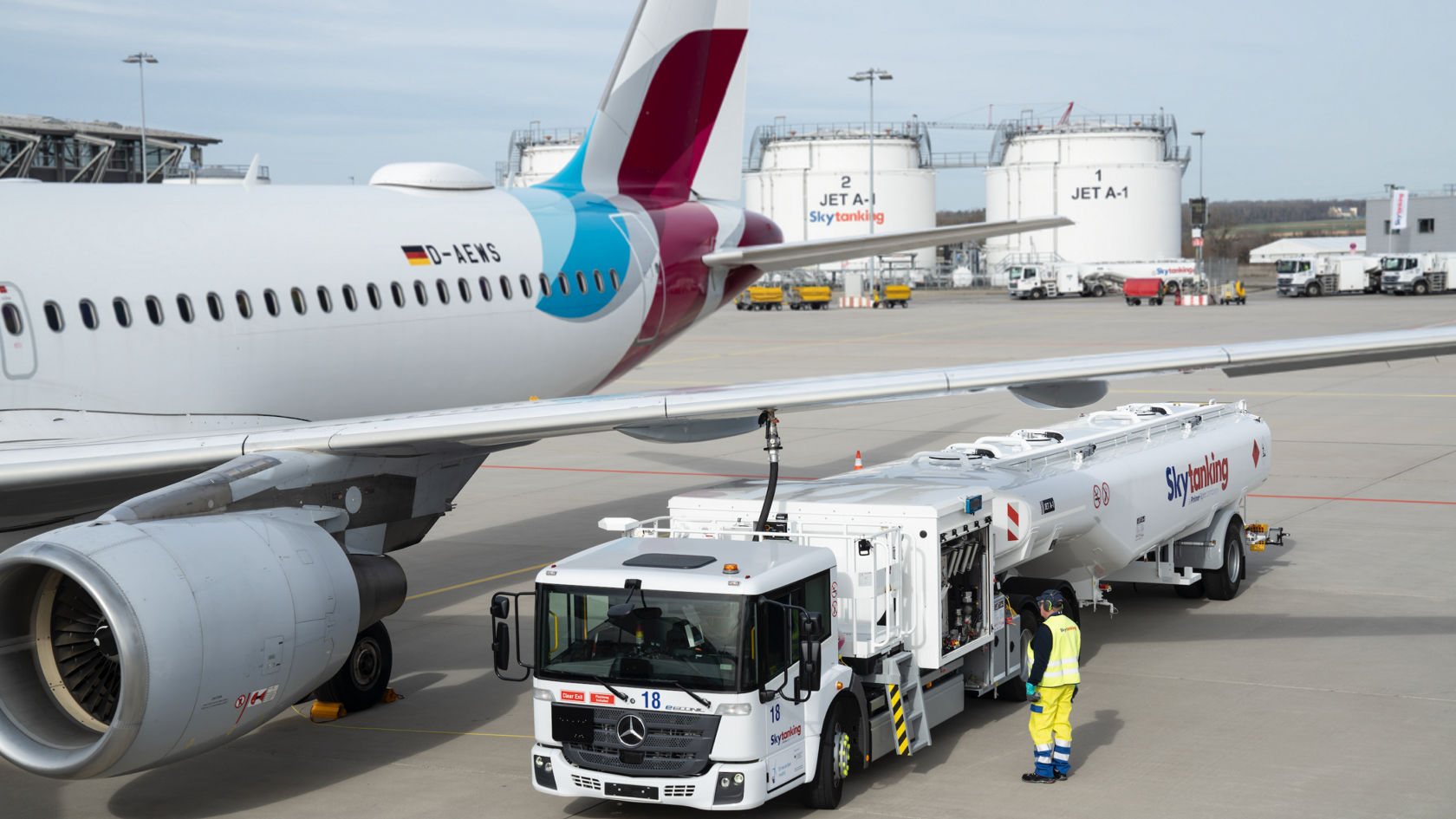  Un Econic blanco, totalmente electrónico, con cisterna, aparcado en la plataforma de un aeropuerto junto a un avión. Un empleado con ropa de alta visibilidad se encuentra junto al vehículo cisterna. En segundo plano se ven otros aviones y sistemas de combustible.