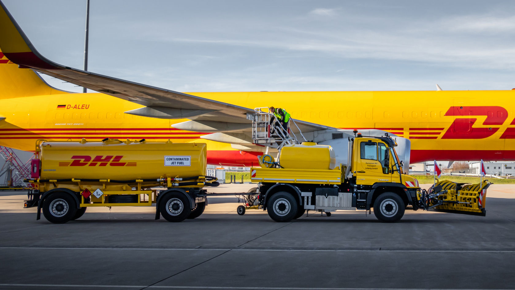 Korjaamonosturilla varustettu keltainen Unimog seisoo keltaisen DHL-lentokoneen vieressä. Huomiovaatteisiin pukeutunut työntekijä on korjaamonosturilla lentokoneen siivellä. Kuorma-auton vieressä on perävaunu, jossa on teksti "Concentrated Jet Fuel".