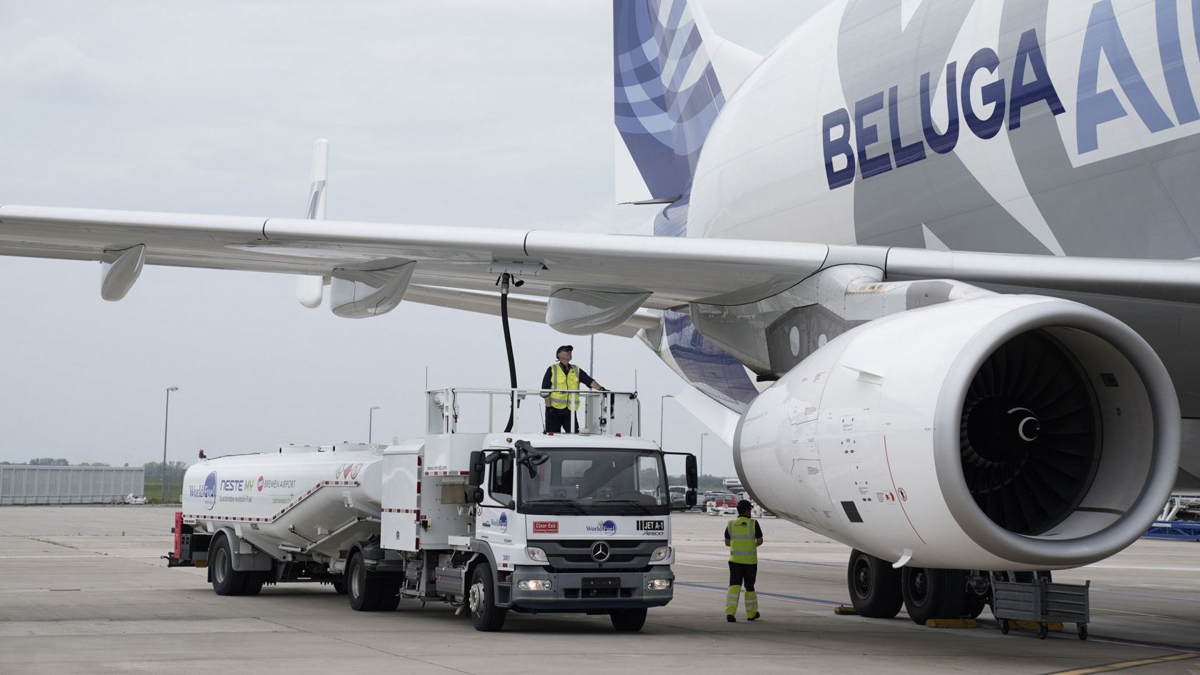 Kuvassa on Airbus Beluga -tyyppinen lentokone, jota tankataan Atego-säiliöautolla. Atego on valkoinen ja siinä on teksti "Neste MY Bremen Airport". Työntekijä seisoo säiliöauton tasanteella tankkaamassa. Toinen työntekijä seisoo lentokoneen moottorin vieressä. Taivas on pilvinen.