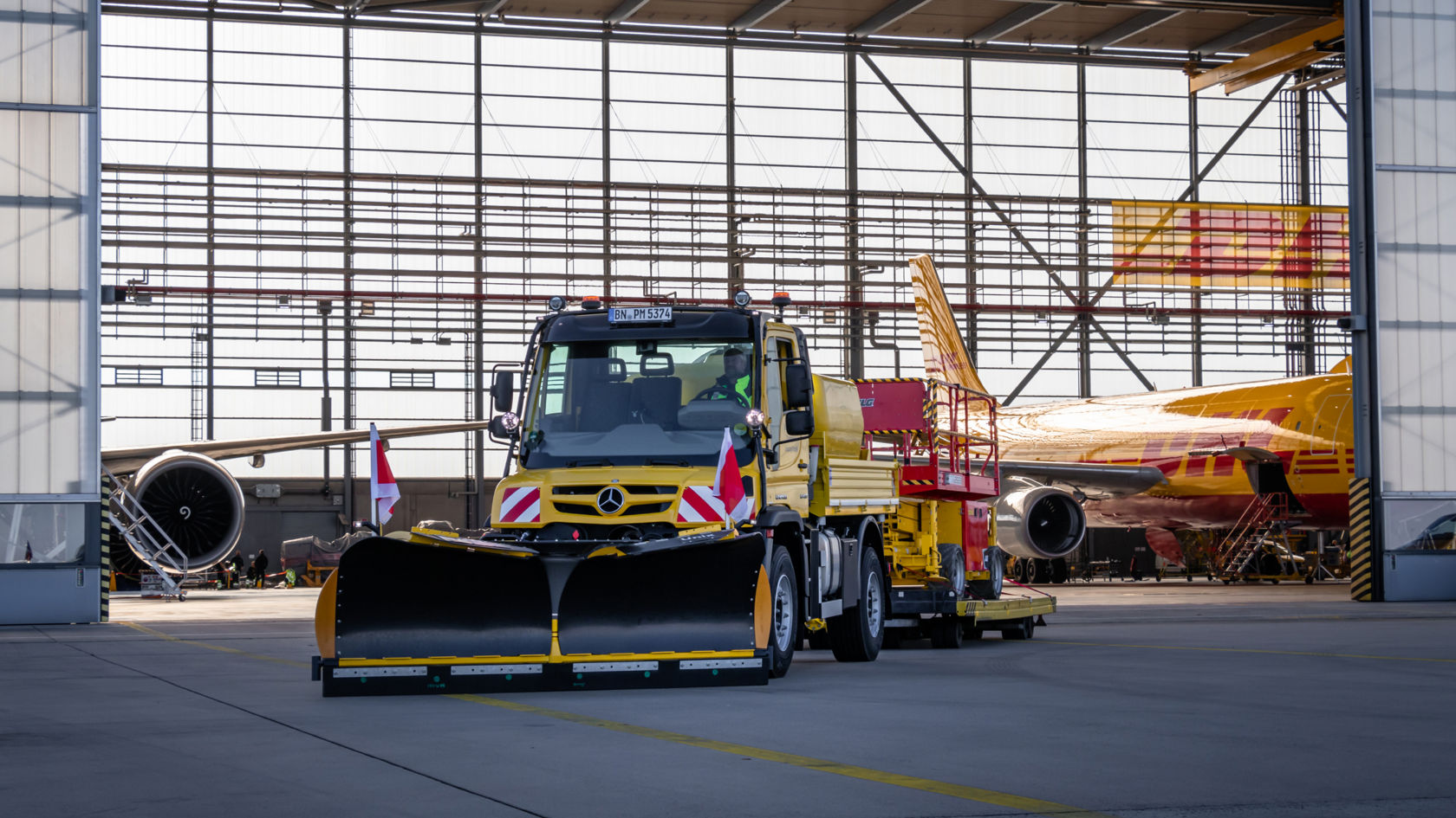 Keltainen Unimog, jossa on lumiaura ja punaisella korjaamonosturilla varustettu perävaunu, seisoo lentokonehallissa keltaisen DHL-lentokoneen edessä. Edessä on kaksi punavalkoista varoituslippua.