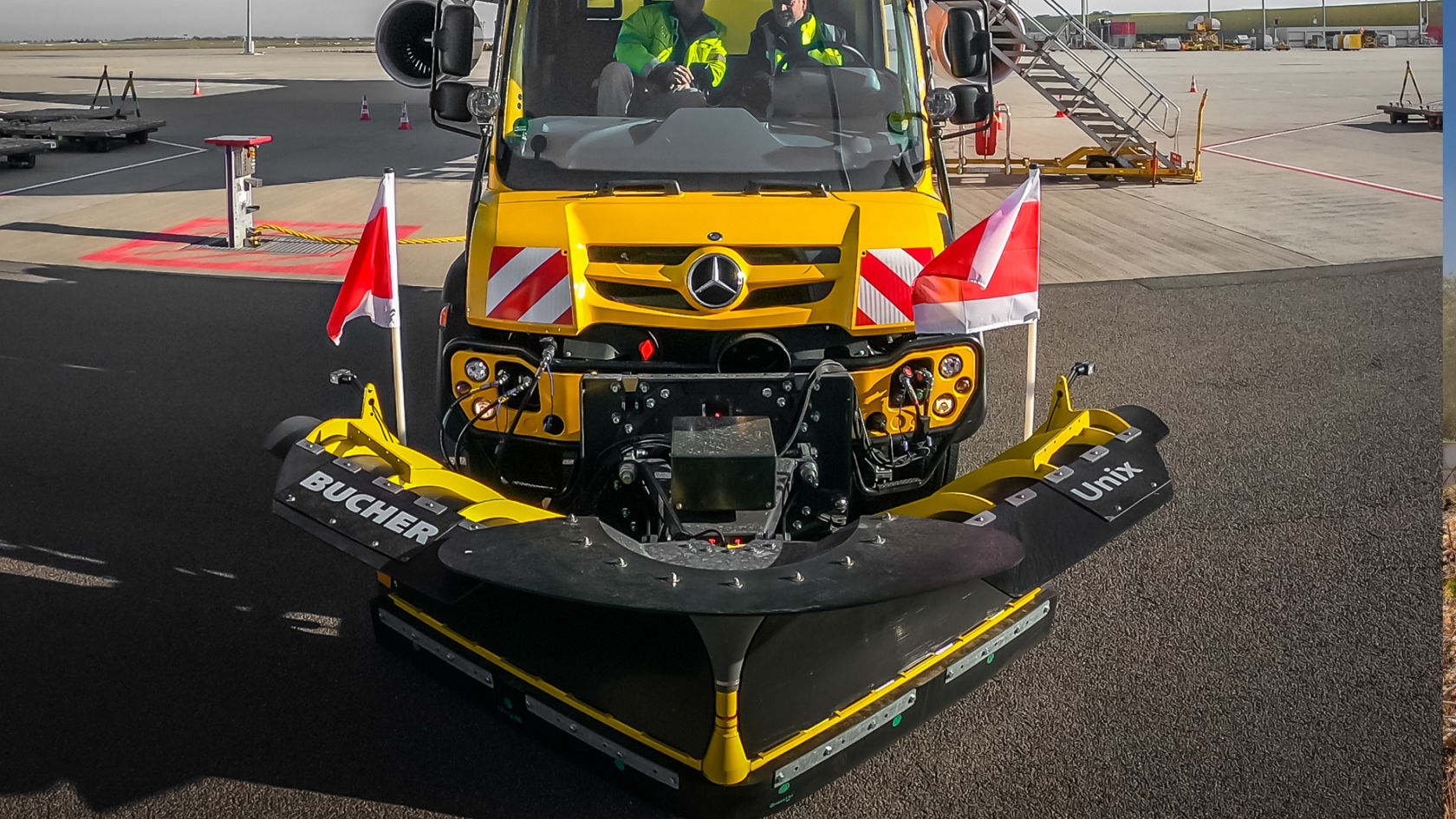 La imagen muestra un primer plano de una cuchilla quitanieves amarilla en un Unimog. La cuchilla quitanieves lleva en los laterales las inscripciones «Bucher» y «Unix». En la cabina del conductor hay dos personas. En los laterales del camión hay colocadas banderas de advertencia rojas y blancas.