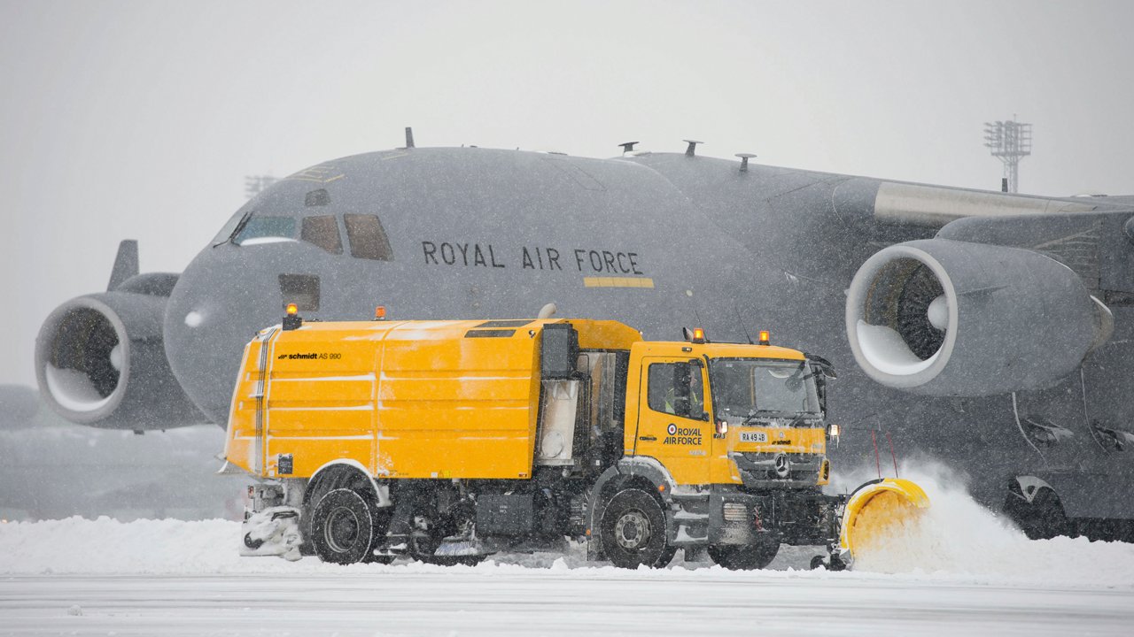 Kuvassa on keltainen Atego-lumiaura, joka auraa lunta kiito- ja laskeutumistieltä. Taustalla on Royal Air Forcen harmaa kuljetuslentokone. Lunta sataa voimakkaasti.