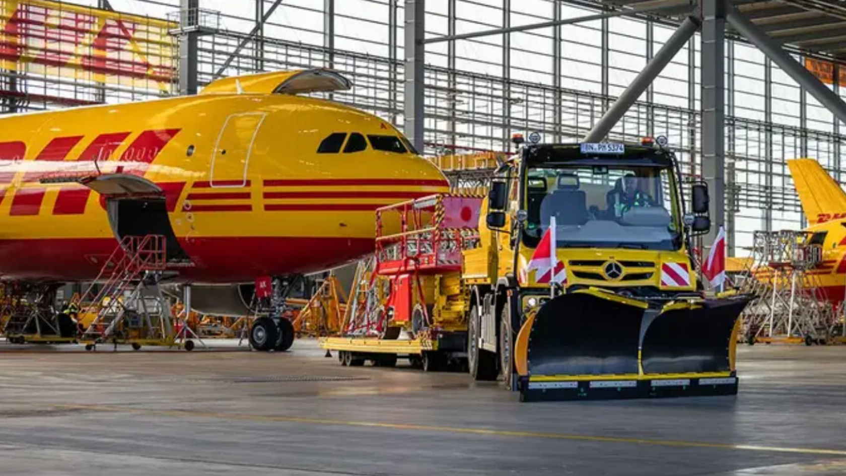 Lentokonehallissa seisoo lumiauralla varustettu Unimog kelta-punaisen DHL-lentokoneen edessä, jota huolletaan. Kuorma-auton takana on korjaamonosturilla varustettu perävaunu, jota todennäköisesti käytetään myös huoltotöissä. Lentokonehalli on suuri ja siinä on teräspalkkirakenne.