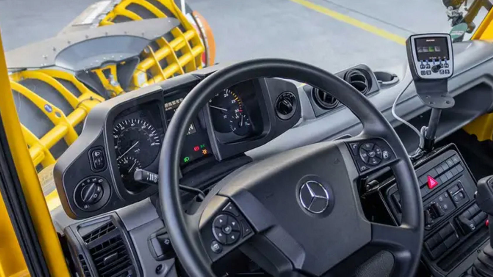 La imagen muestra un primer plano del tablero de instrumentos de un camión. Se muestran el volante, los indicadores de velocidad, número de revoluciones y contenido del depósito, así como diversos elementos de mando. A la derecha del volante se encuentra un pequeño visualizador.