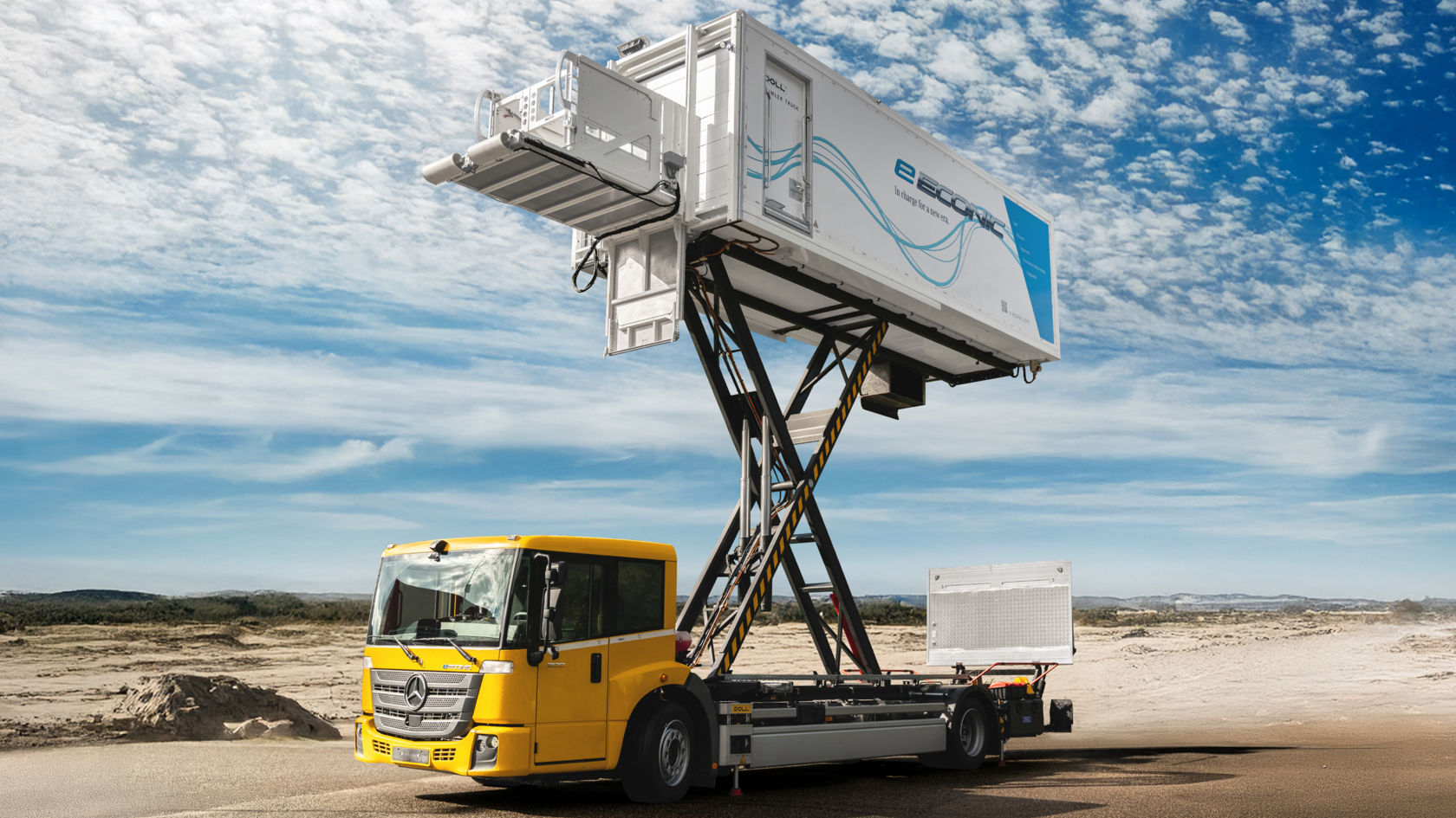 Un Econic amarillo con una trampilla de carga grande plegada está aparcado sobre un terreno arenoso o sin asfaltar. En la superestructura de la trampilla de carga se lee «e econic», lo que indica una versión totalmente electrónica. La trampilla de carga está muy extendida, de modo que la superestructura parece un contenedor. En el fondo se ve un cielo azul con algunas nubes.