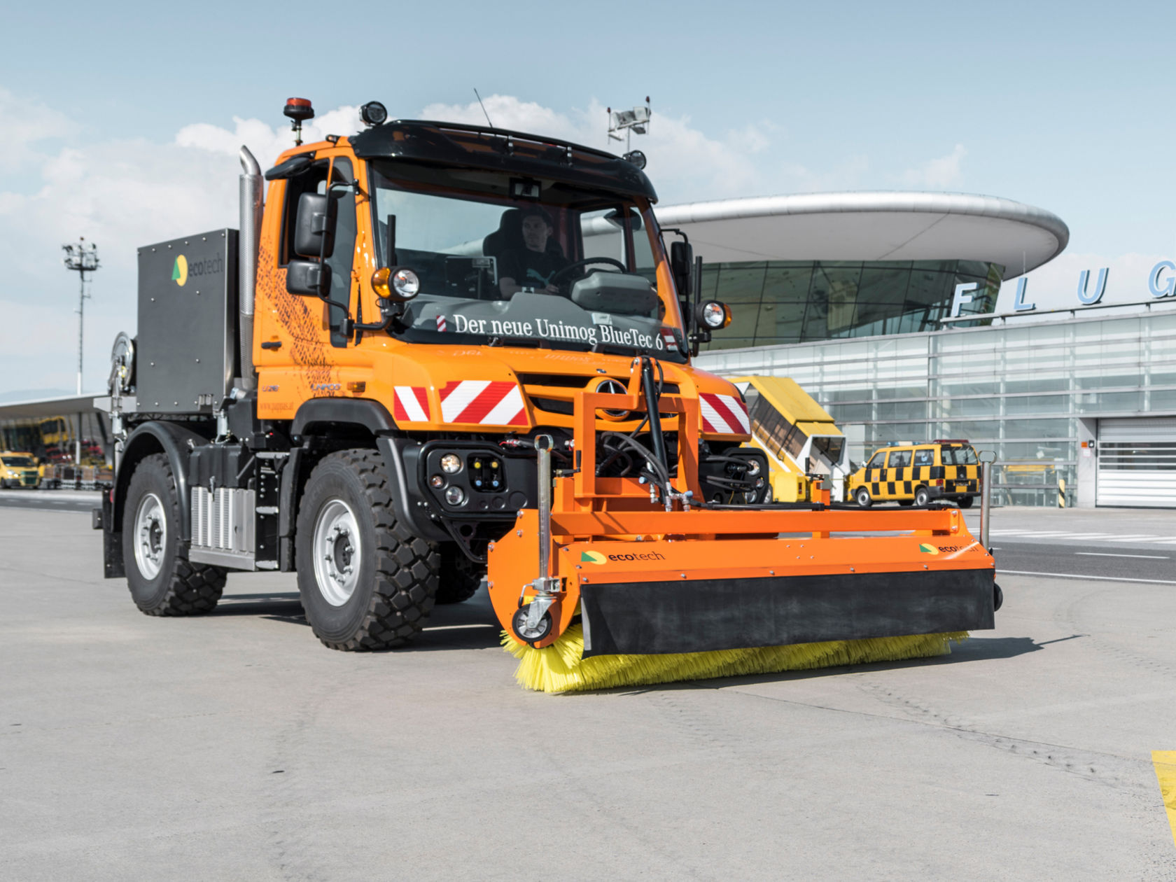 Oranssi Unimog, jonka edessä on lakaisukone, ajaa lentokentän asematason yli. Ohjaamossa lukee "Der neue Unimog BlueTec 6". Taustalla näkyy muita lentokoneita ja lentokenttärakennus.