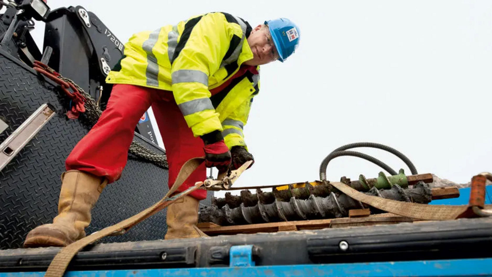 Een arbeider in een gele veiligheidsjas en een blauwe helm zekert met een spanband een groot mechanisch onderdeel op een laadplatform van een vrachtwagen.