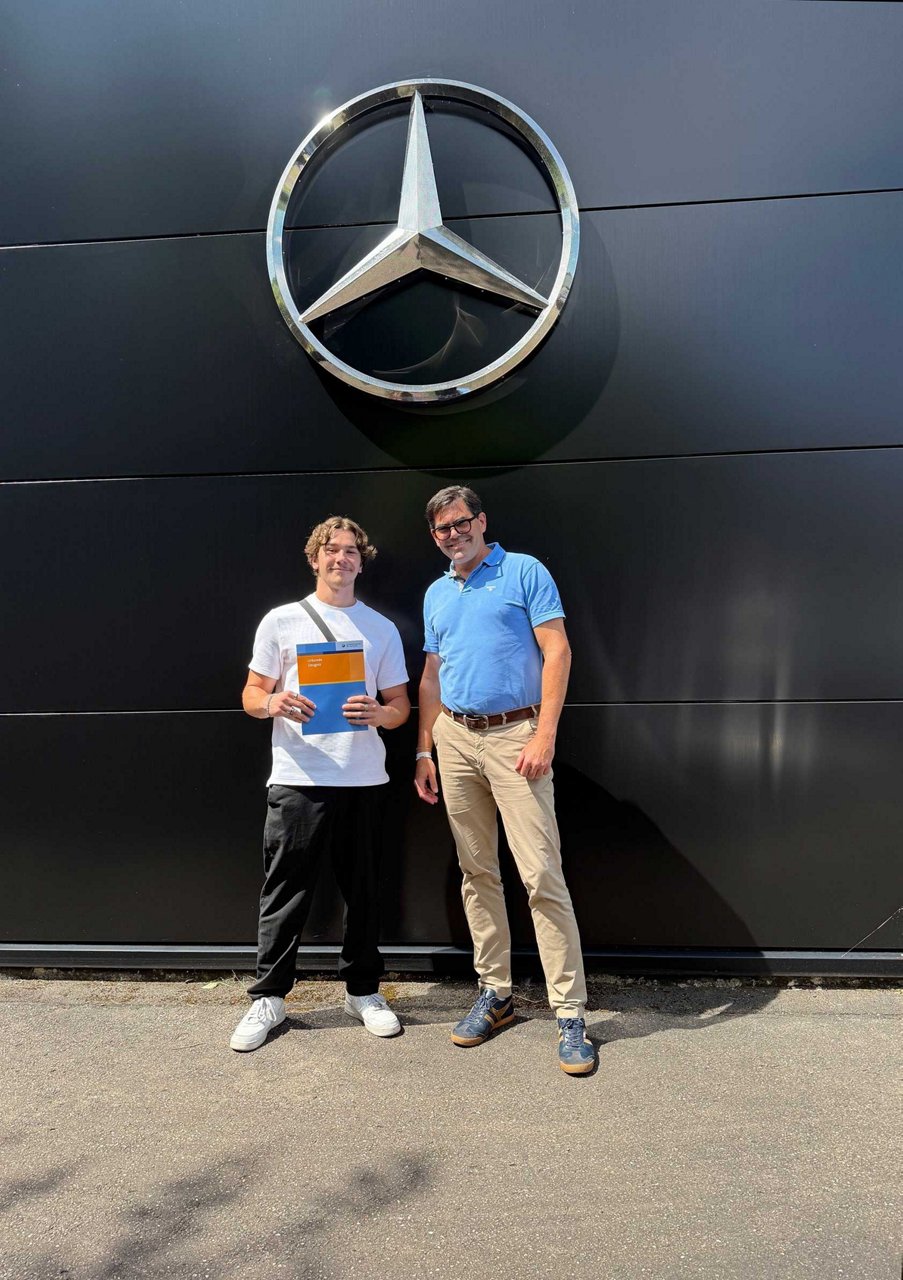 zwei Personen stehen vor einer schwarzen Wand mit einem großen Mercedes Stern, eine hält ein Dokument in Händen.