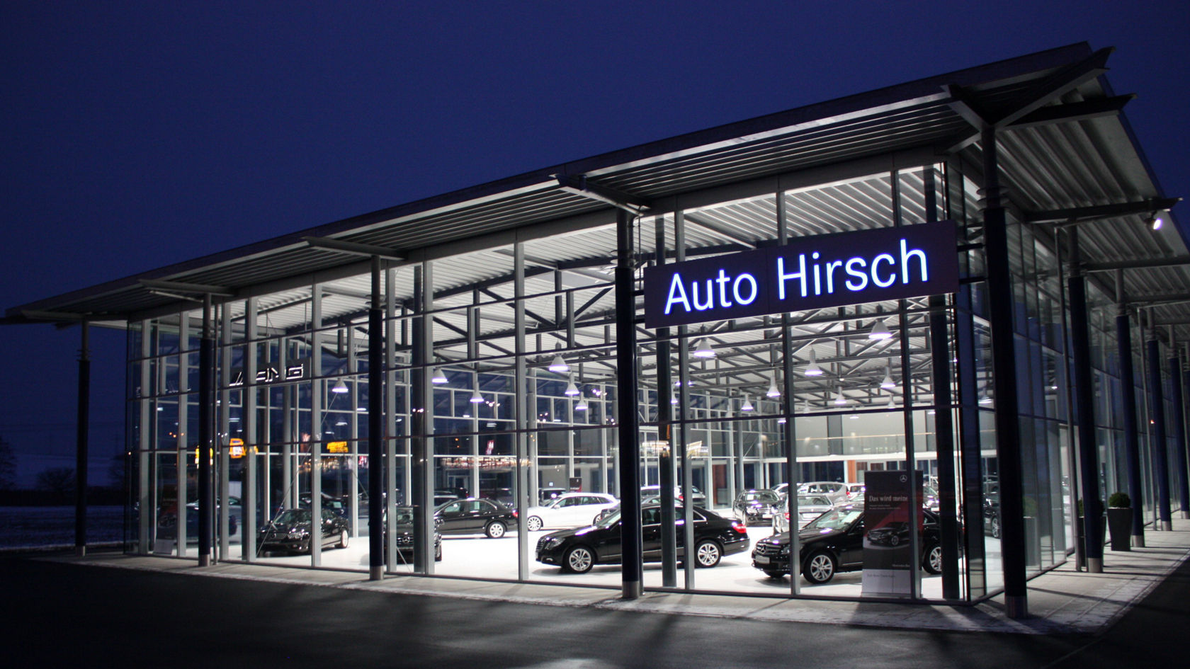 Außenansicht des Autohauses Hirsch in Arnstorf bei Nacht. Das moderne Gebäude mit Glasfassade ist hell erleuchtet, im Inneren sind mehrere Fahrzeuge zu sehen. Über dem Eingang ist der Schriftzug „Auto Hirsch“ sichtbar.