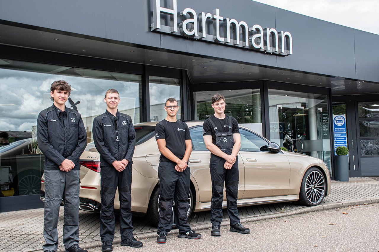 Vier Auszubildende des Mercedes-Benz Autohauses Hartmann in Lahr stehen in schwarzer Arbeitskleidung vor dem Autohaus. Neben ihnen ist ein beiges Fahrzeug geparkt. Über dem Eingang ist das Firmenschild „Hartmann“ sichtbar.