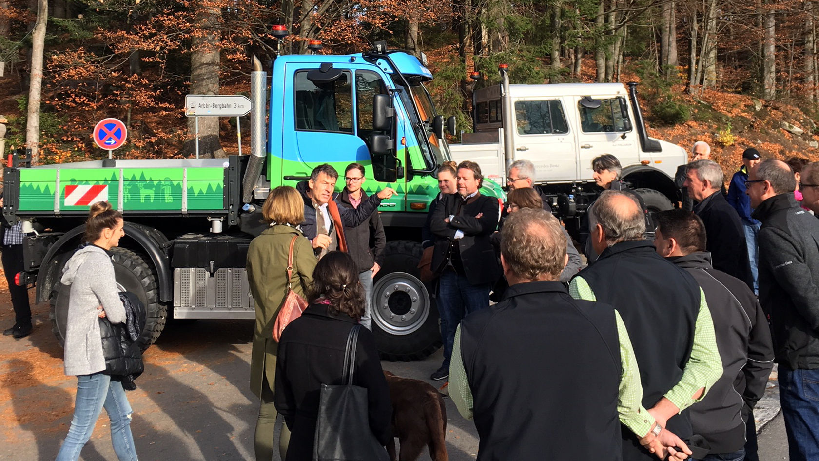 A group of people is gathered around a Mercedes-Benz Unimog as a speaker provides information about the vehicle and its functions, surrounded by an autumnal woodland landscape.