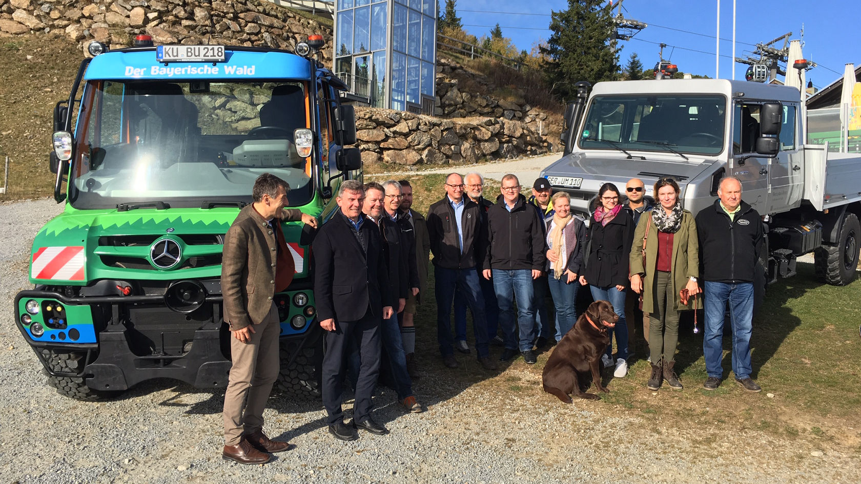 Two Mercedes-Benz commercial vehicles stand beside a group of people gathered on a meadow, while a dog lies nearby.