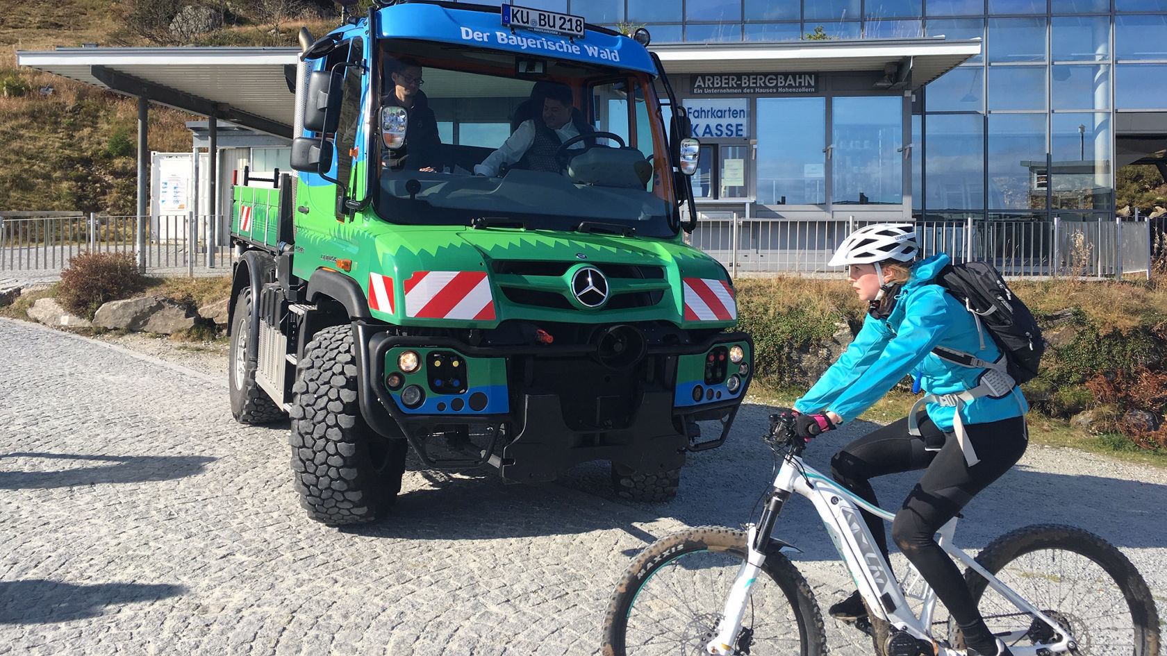 A green Mercedes-Benz off-road vehicle stands at a mountain station, while a cyclist in a blue jacket and a white helmet rides past.