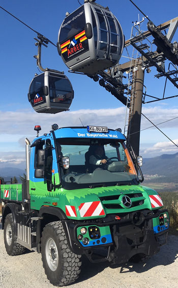A green Mercedes-Benz Unimog vehicle stands beneath a cable car carrying gondolas over a mountain landscape. A clear sky completes the scene.