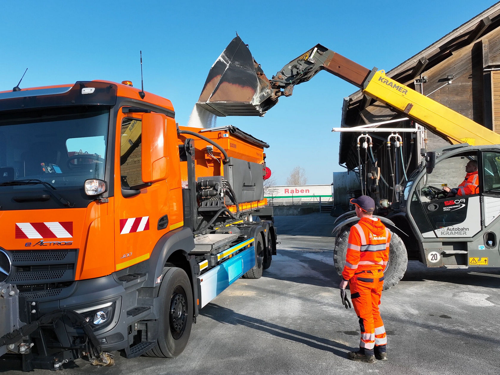 Ein orangefarbener eActros von Mercedes-Benz steht an einer Baustelle, während eine hydraulische Ladebühne Material von einem Bagger auflädt. Ein Arbeiter in orangefarbener Sicherheitskleidung überwacht den Prozess.
