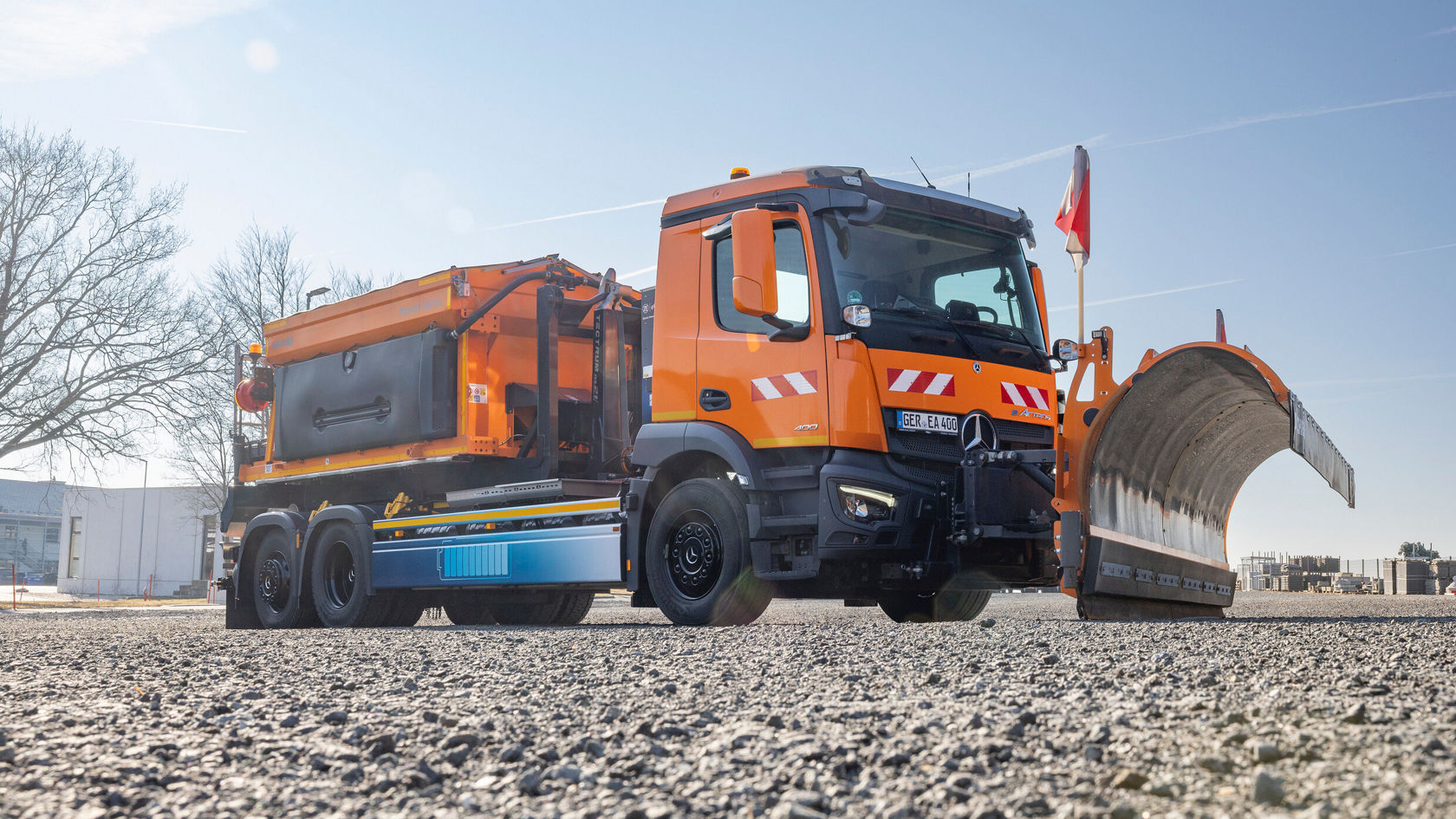 Ein Mercedes-Benz eActros in leuchtendem Orange steht auf einem Kiesplatz. Er ist mit einem Schneepflug ausgestattet.
