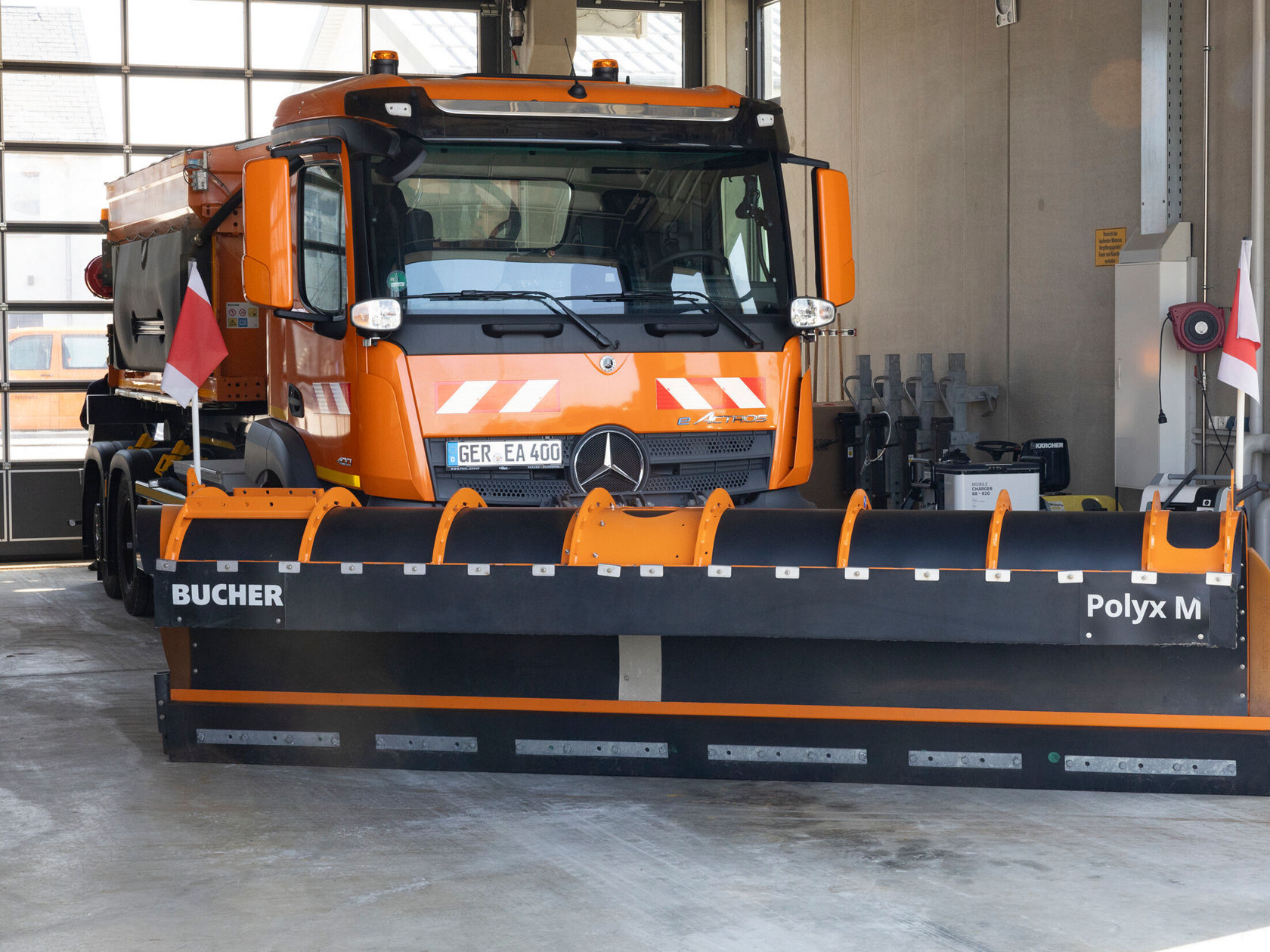 Ein orangefarbener Mercedes-Benz eActros mit einer Schneeräumschaufel vorne steht in einer Garage. Die Schaufel ist mit "BUCHER Polyx M" gekennzeichnet.