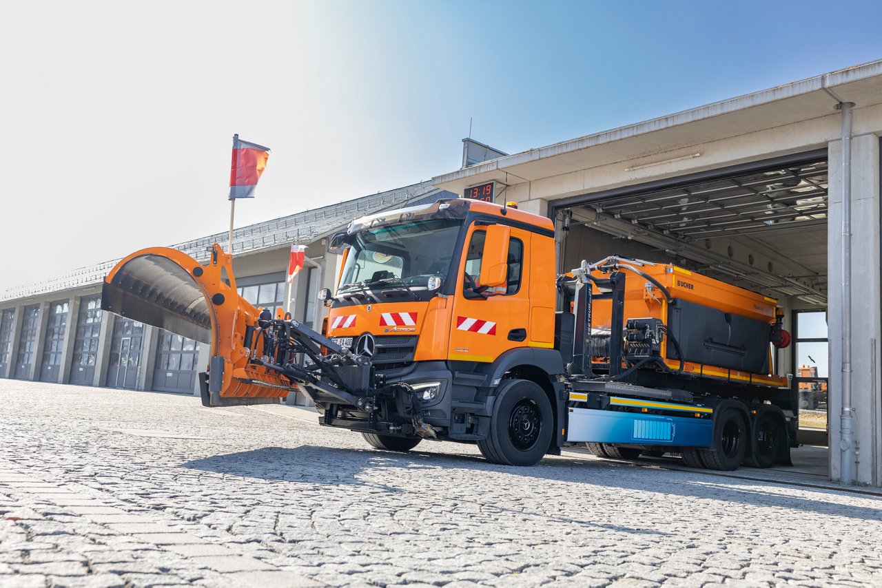Ein Mercedes-Benz eActros Lkw mit einem vorderen Schneepflug fährt auf einer Autobahn. Der Lkw ist orange und trägt Warnflaggen. 