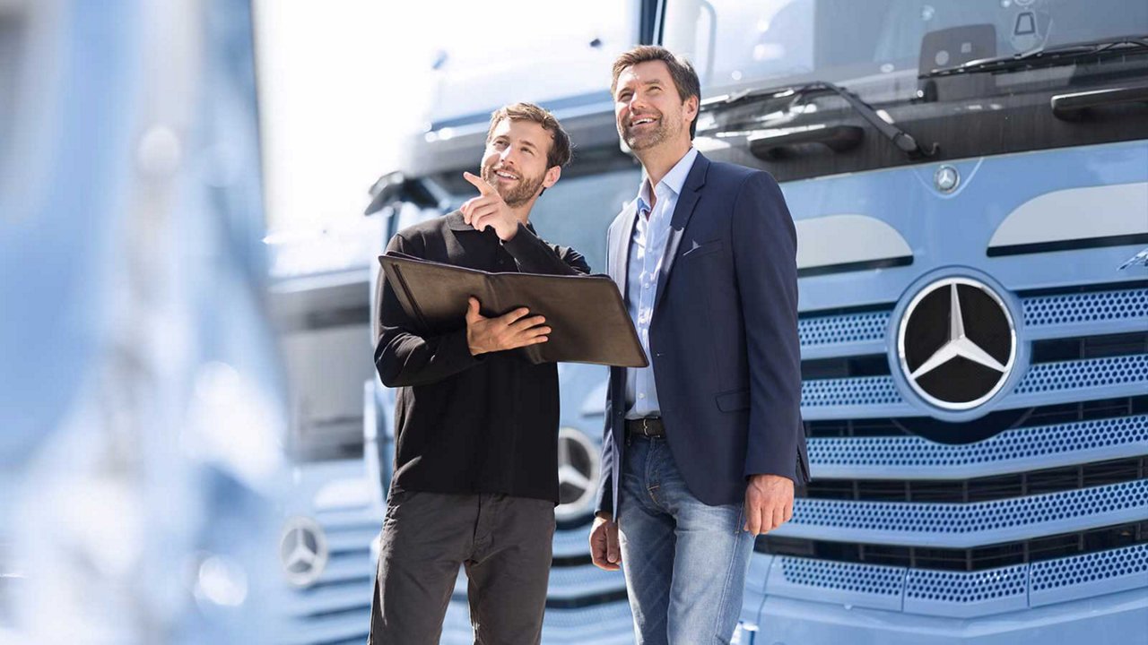 Zwei Männer stehen vor einem blauen Lkw mit Mercedes-Benz-Logo. Eine Person hält eine Mappe und zeigt auf einen Punkt außerhalb des Bildes, während die andere aufmerksam schaut.