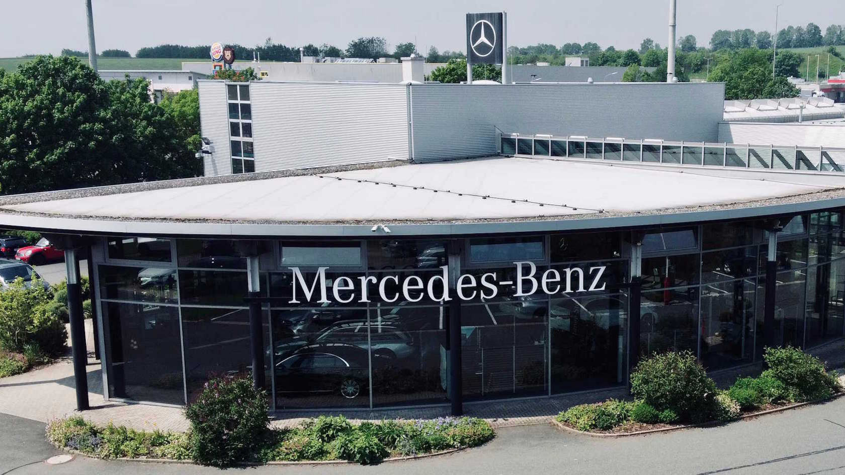 Außenansicht des Mercedes-Benz Autohauses Hahn in Münchberg. Das Gebäude hat eine runde Glasfassade mit flachem Dach und ist von gepflegten Grünflächen und Sträuchern umgeben. Auf dem Dach ist das Mercedes-Benz Logo auf einem Schild sichtbar.