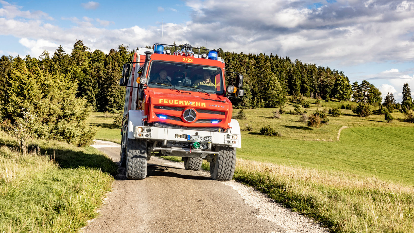 Ein Feuerwehrfahrzeug der Marke Mercedes-Benz fährt auf einer ländlichen Straße, umgeben von Wiesen und Bäumen unter blauem Himmel.