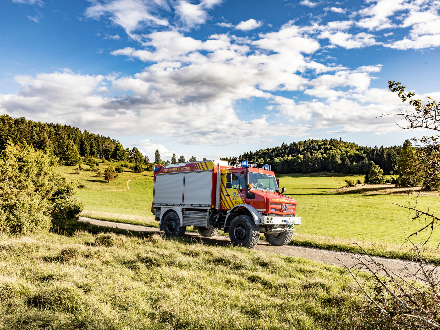Ein roter Mercedes-Benz Feuerwehrwagen fährt auf einer kurvenreichen Landstraße durch eine malerische, grüne Landschaft mit Bäumen und einem weiten Himmel.