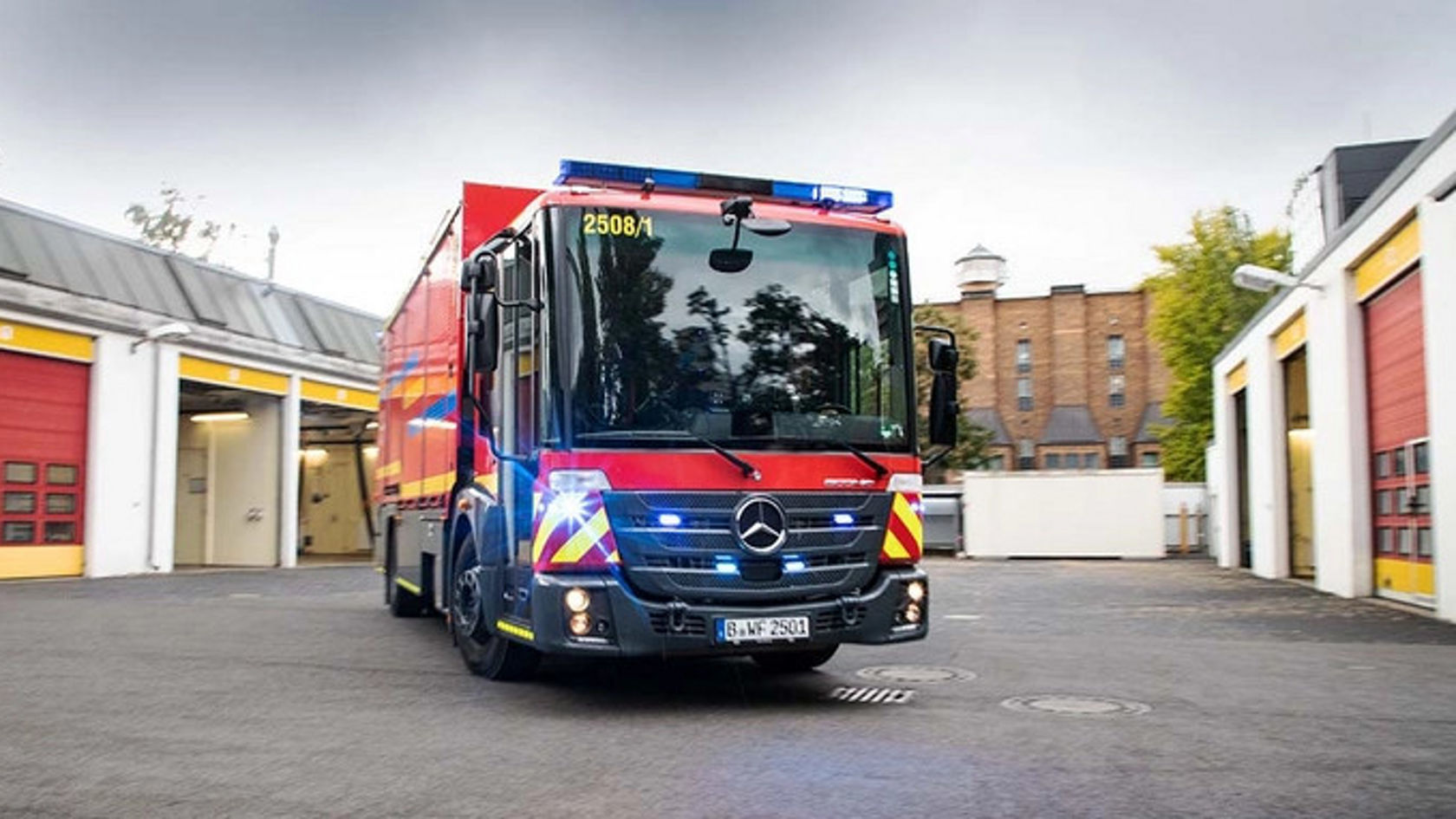 Ein Mercedes-Benz Feuerwehrauto fährt aus einer Feuerwache. Das Fahrzeug ist rot mit blauen Lichtern und reflektierenden Streifen, umgeben von Garagentoren.
