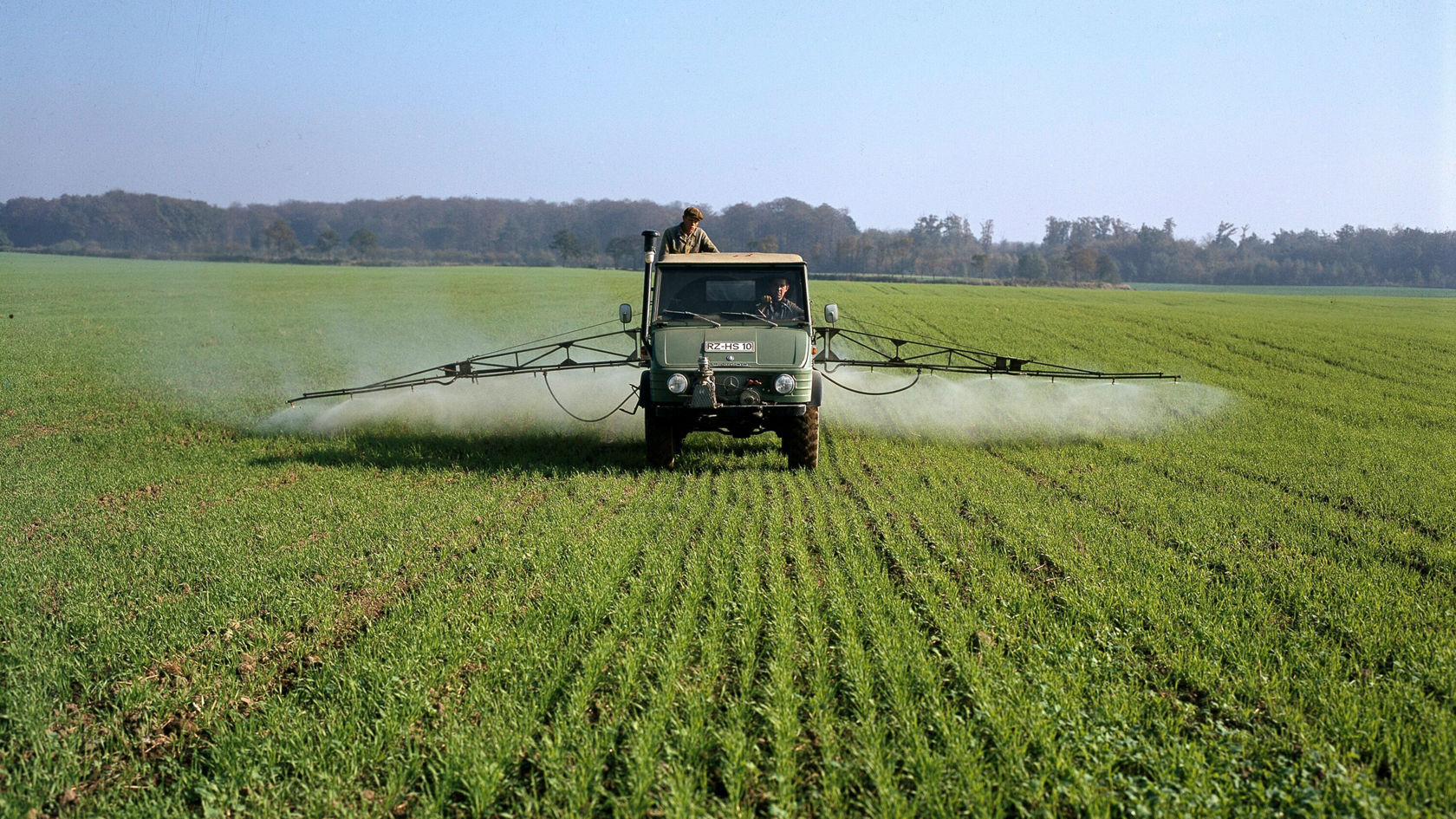 An Unimog from Mercedes‑Benz sprays fertilizer onto a field with green vegetation. The machine distributes a liquid evenly as it drives through the rows.