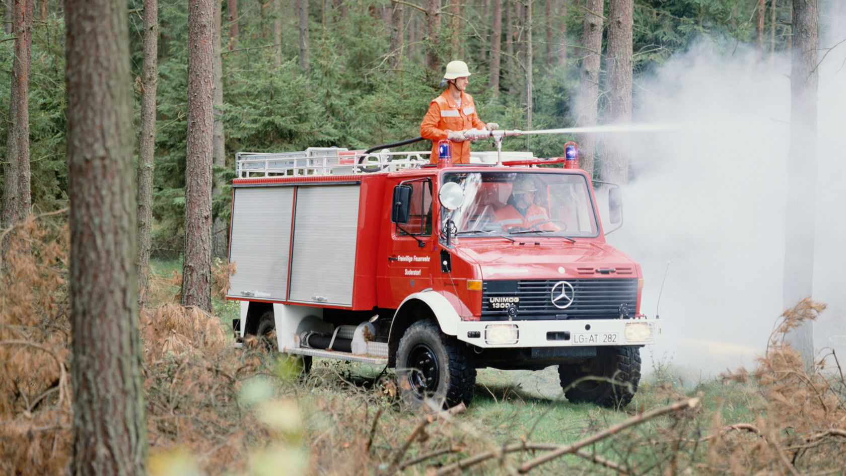 A red Mercedes-Benz Unimog fire truck is standing in the forest while a firefighter on top operates a hose to extinguish smoke.