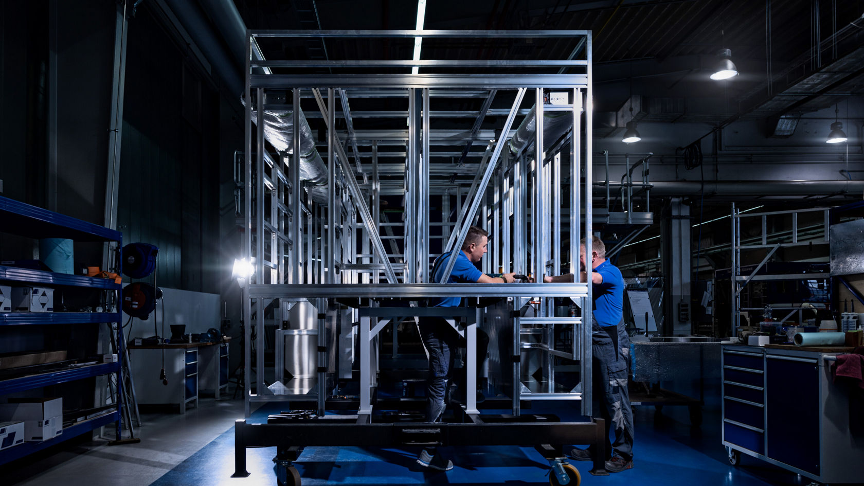 Deux ouvriers en T-shirts bleus assemblent une construction métallique structurée dans un hall industriel, entourés d'outils et de machines.